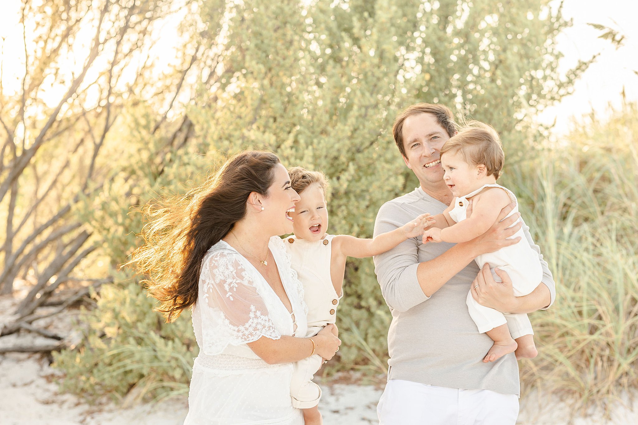 A happy family of 4 laughs and plays with 2 toddlers on a windy beach at sunset while visiting kid friendly hotels miami