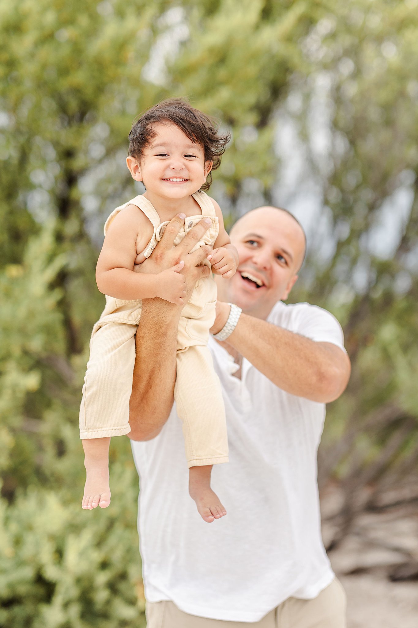 A smiling toddler boy is lifted by dad while playing on a beach