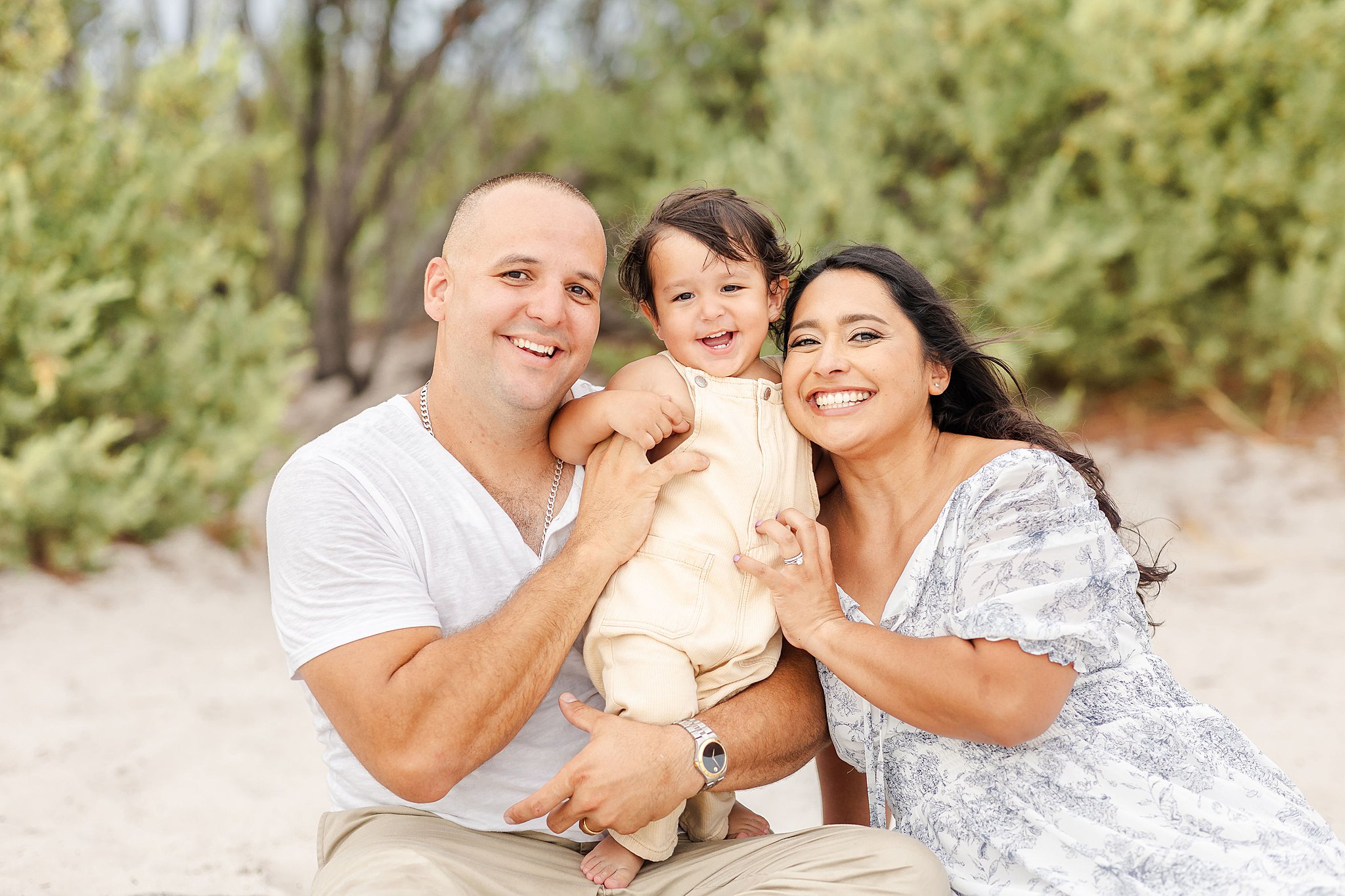 A toddler in a yellow onesie stands in dad's lap on a beach while hugging mom and laughing before visiting kid friendly restaurants miami