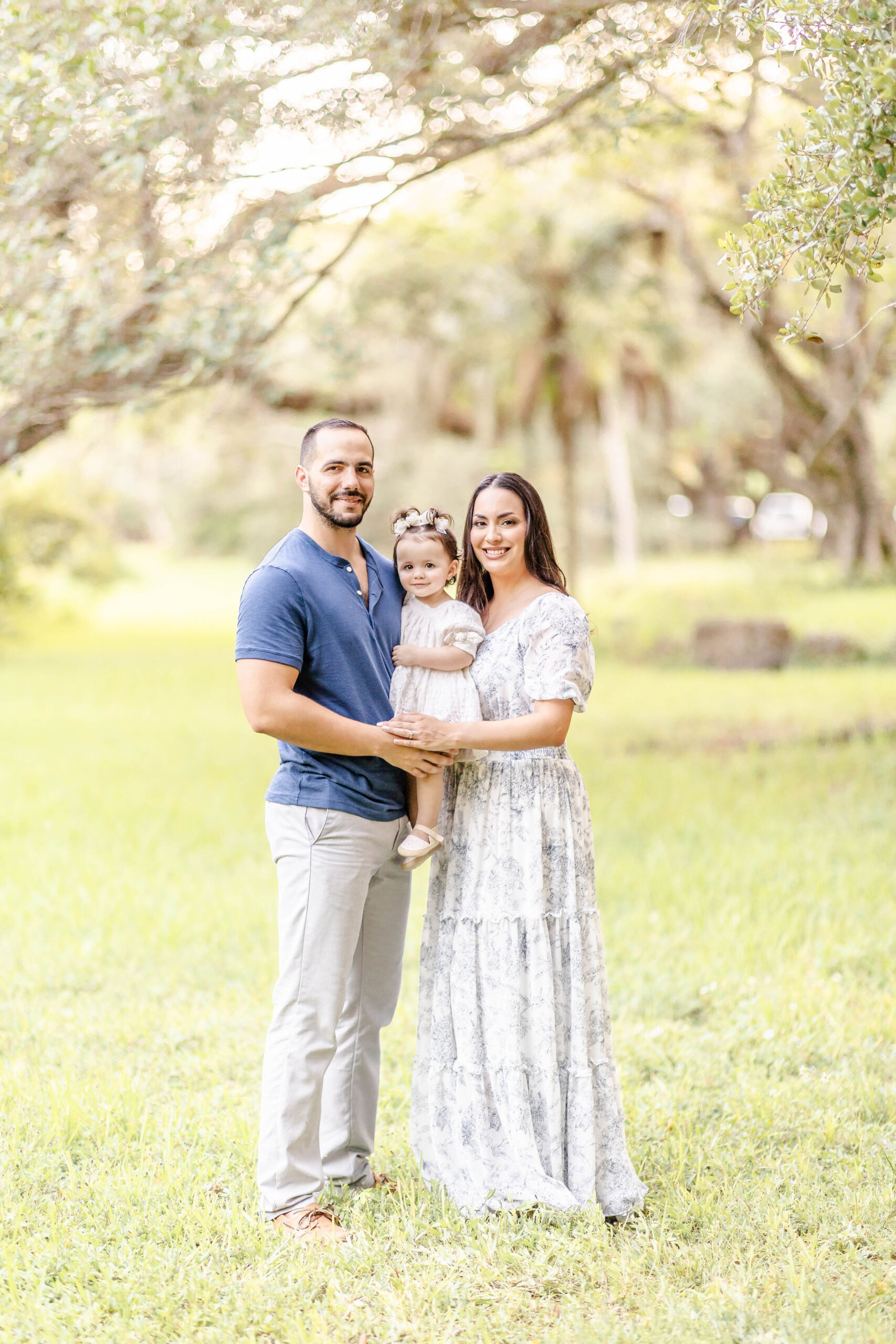 A happy mom and dad stand in a park holding their smiling toddler daughter between them