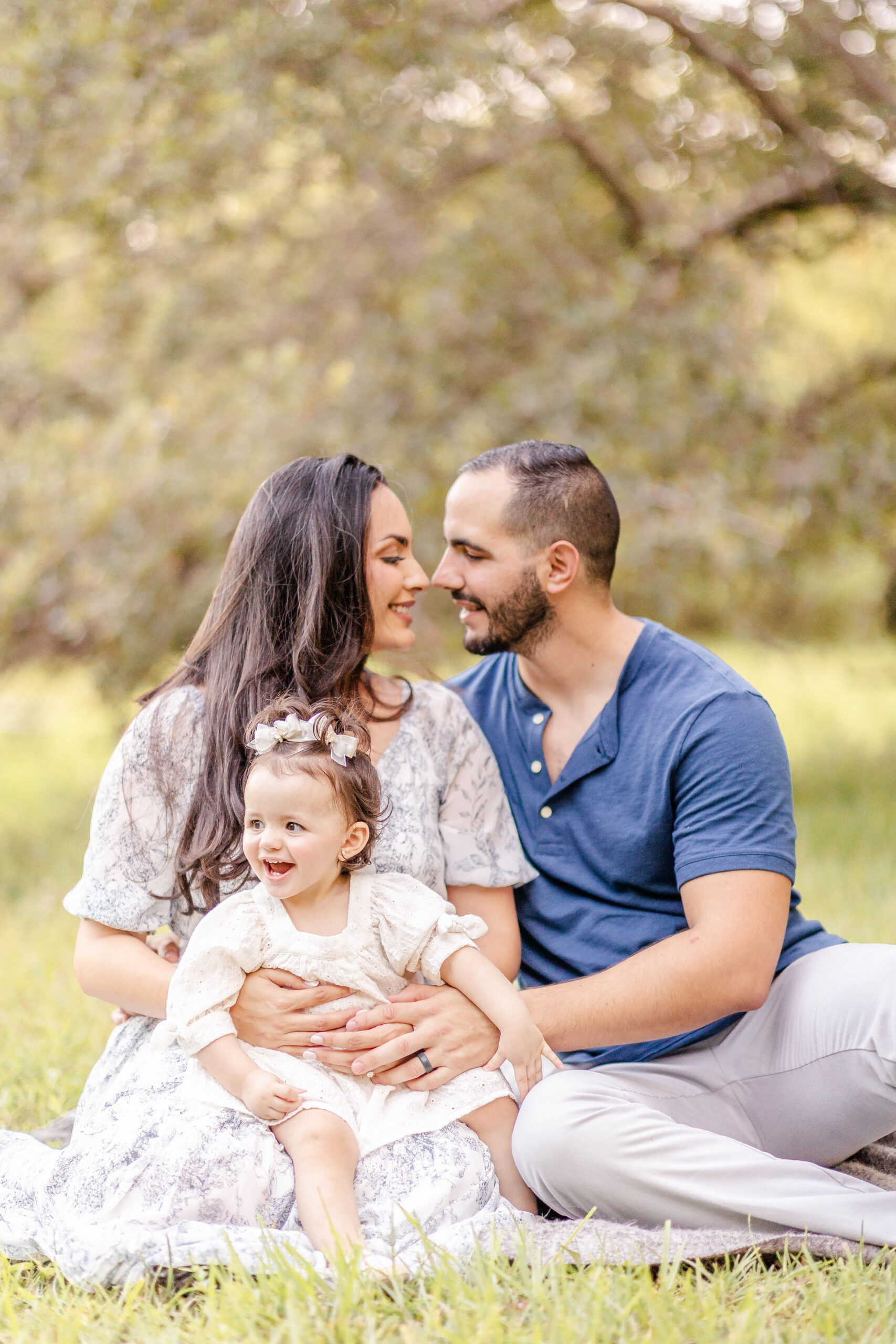 Happy parents lean in for a kiss while their toddler daughter laughs in moms lap in a park before visiting private preschools in miami