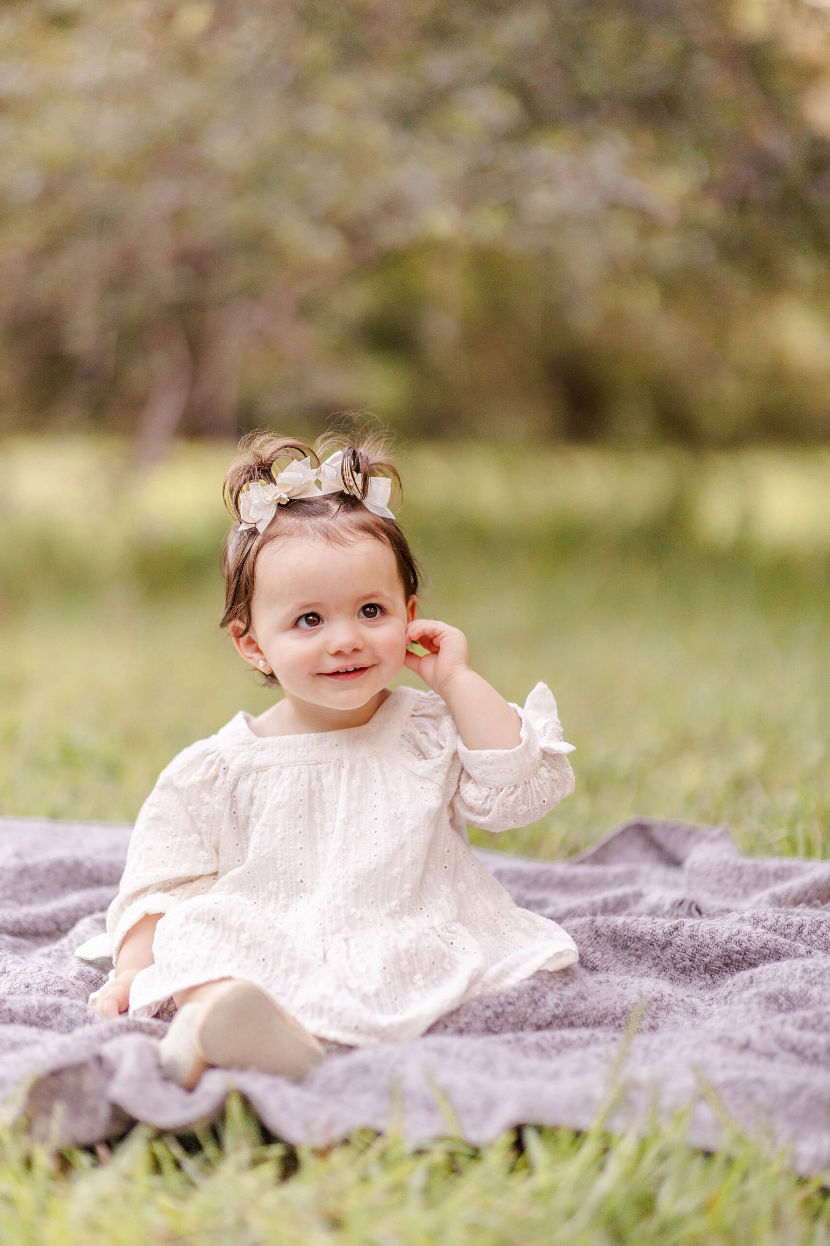 A toddler girl sits on a picnic blanket in a white dress in a park before attending private preschools in miami