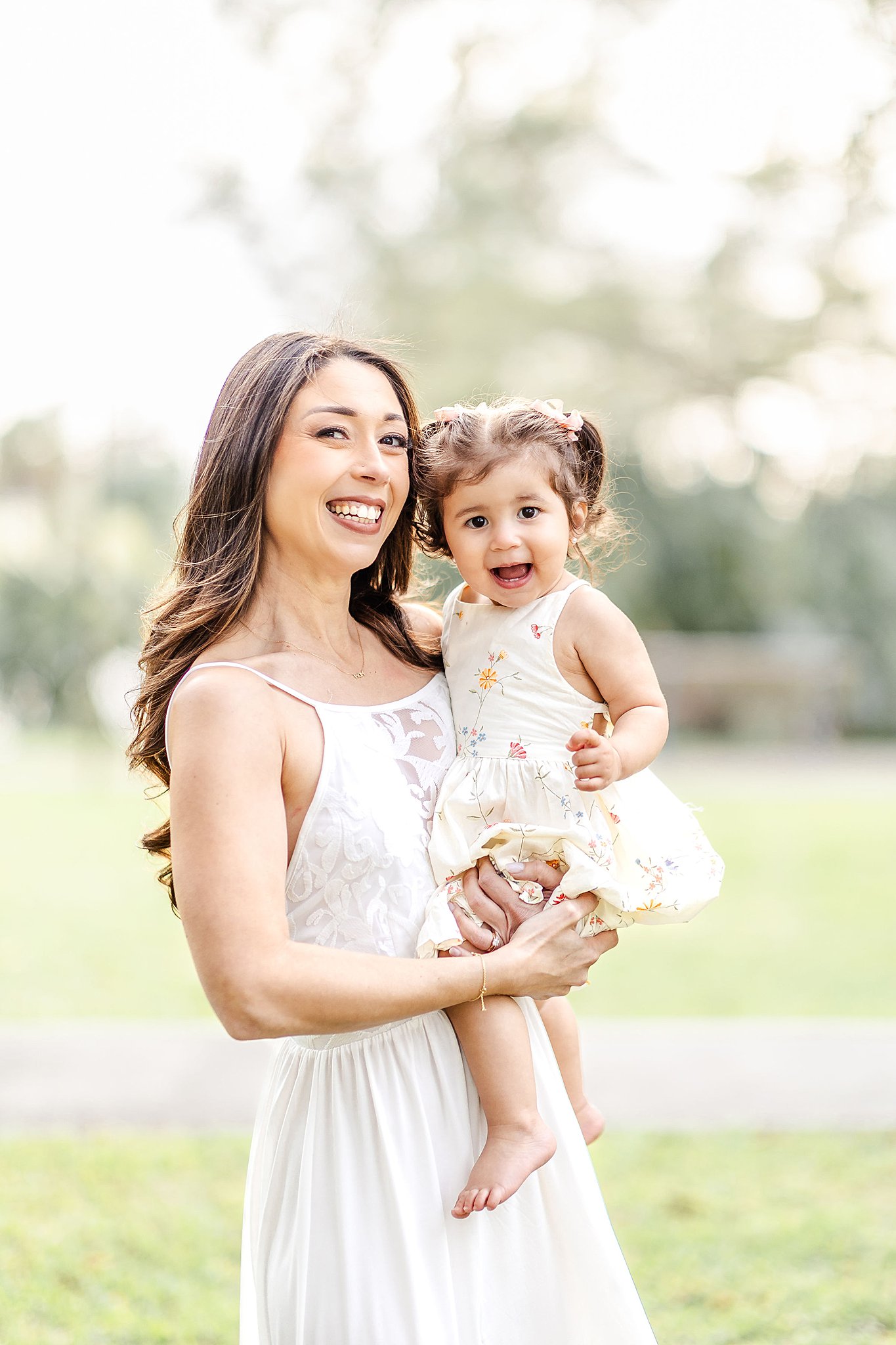 A happy mom and toddler daughter in white dresses stand in a park field smiling and giggling