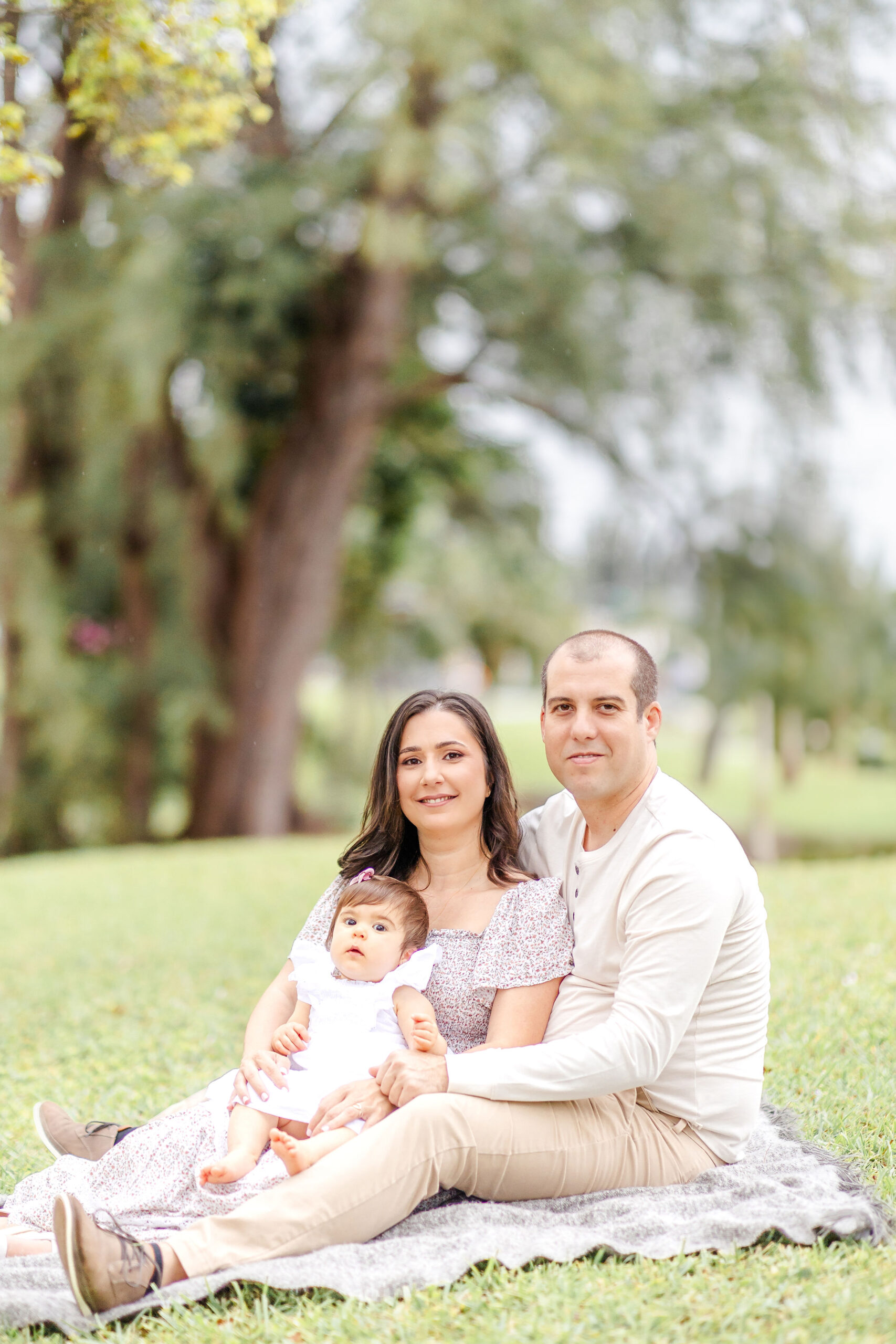 A toddler girl sits in mom and dad's lap on a blanket in a park lawn