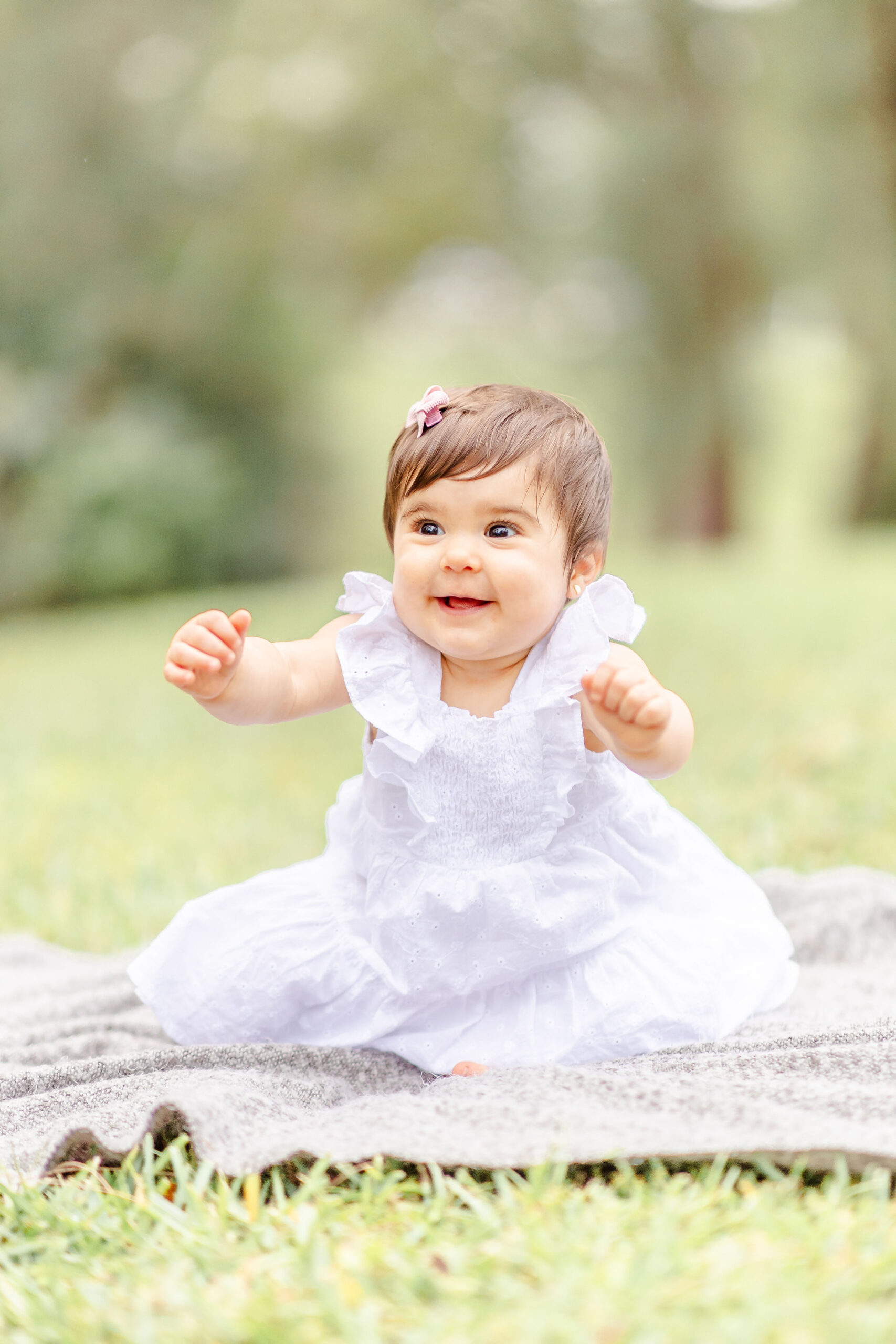 A young toddler girl in a white dress reaches out while sitting on a blanket in a park before visiting splash pads miami