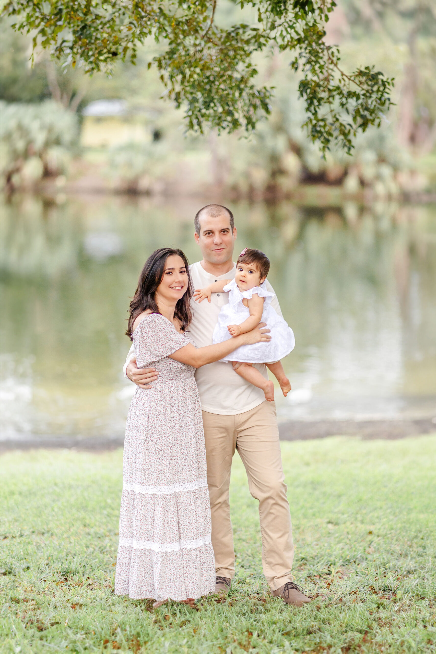 A mom and dad stand under a tree by a lake while holding their toddler daughter before visiting splash pads miami