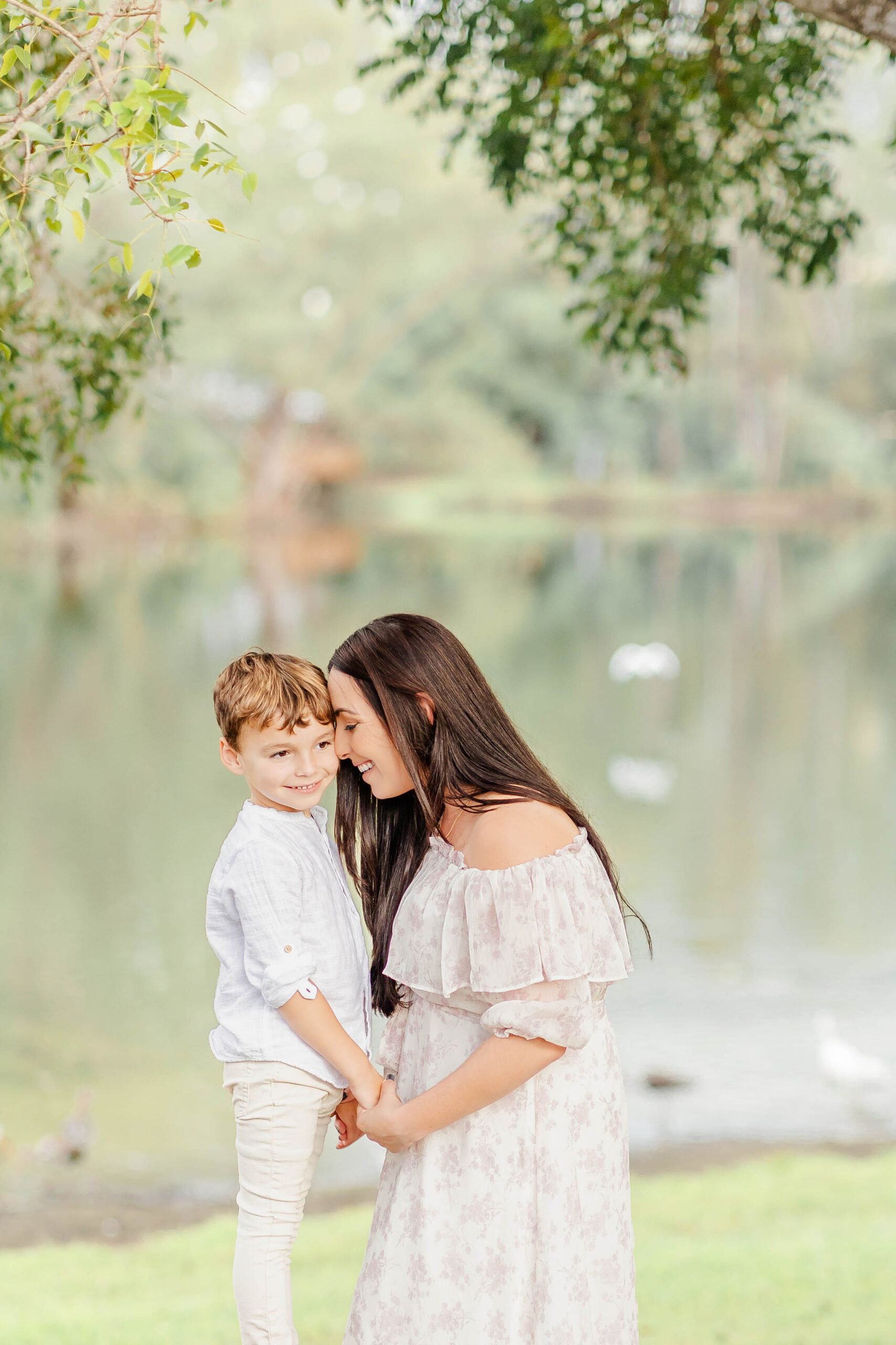 A mom snuggles and holds hands with her toddler son in a park by a lake