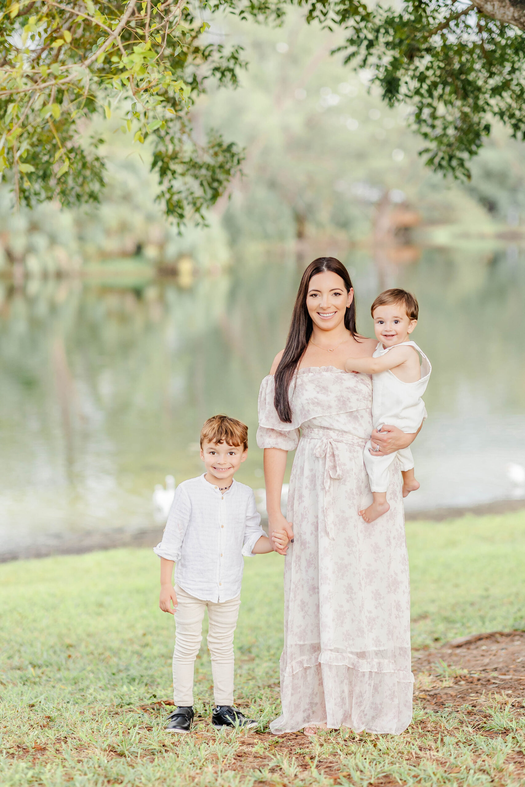 A happy mother stands on a lake edge holding her toddler son and holding hands with her older son before visiting trampoline parks miami