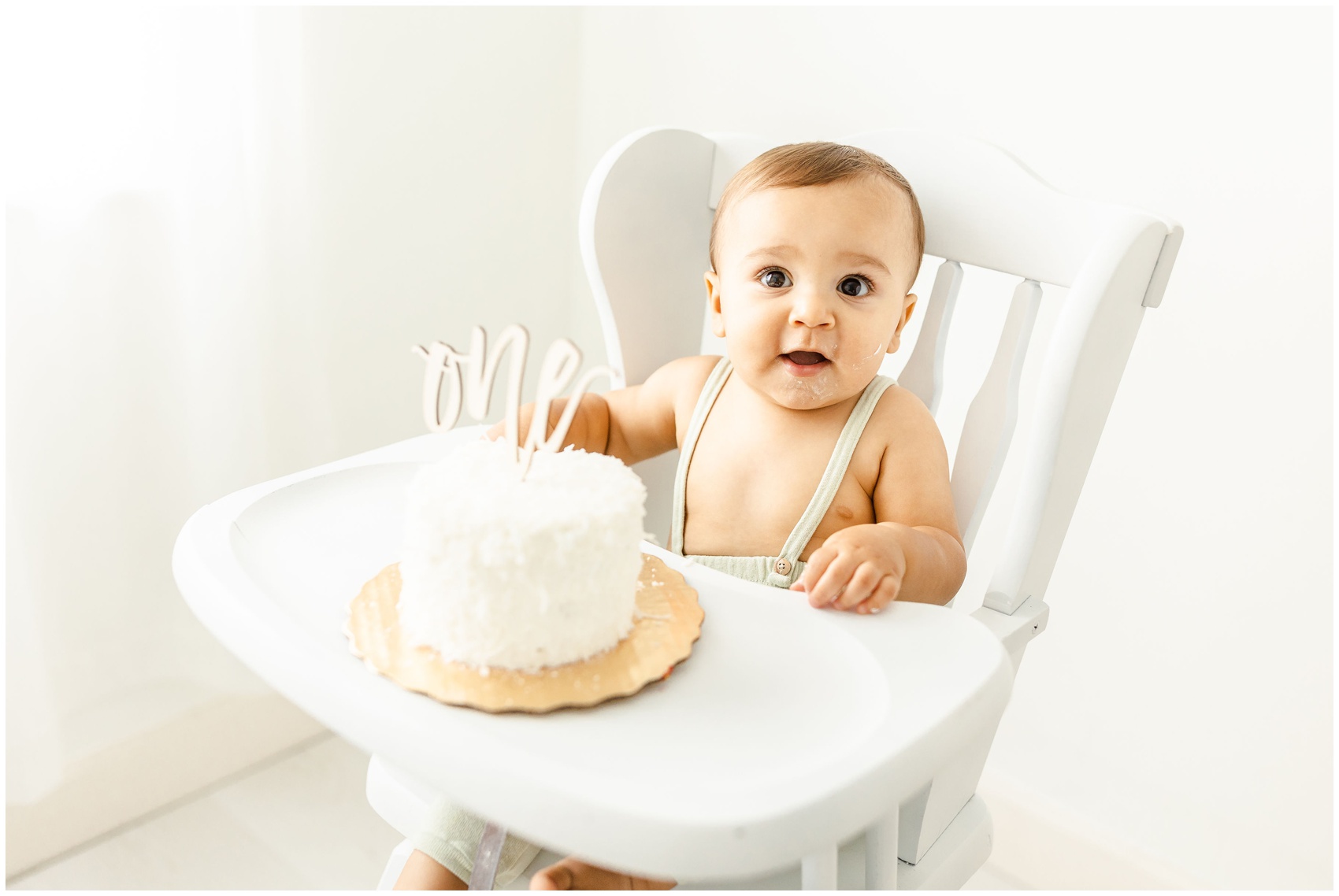 A surprised toddler boy puts his hand in his birthday cake while in a white high chair in green overalls