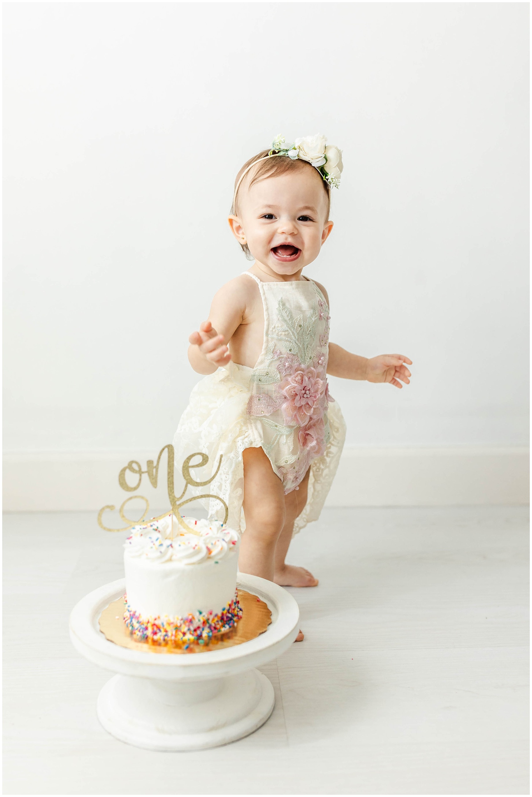 A happy toddler girl walks in a white dress behind her first birthday cake