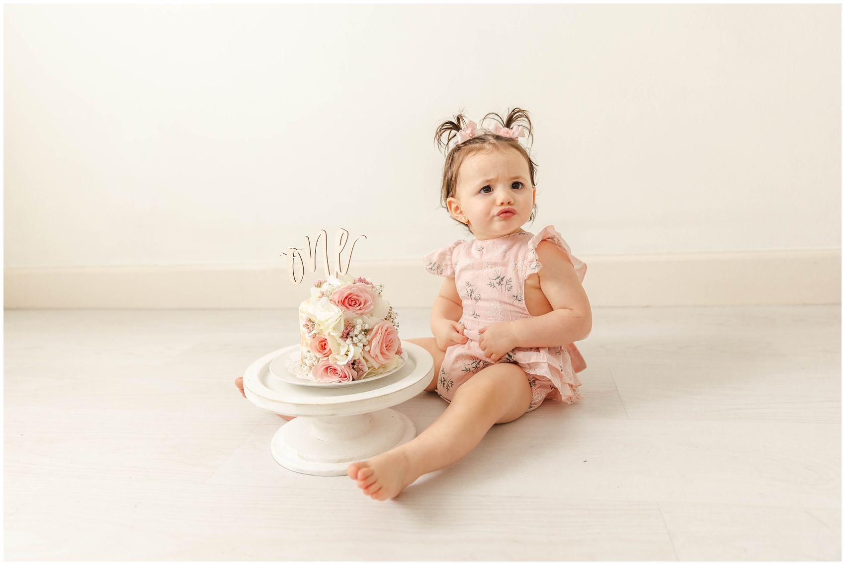 A young toddler girl in a pink onesie sits with her flower covered cake in a studio