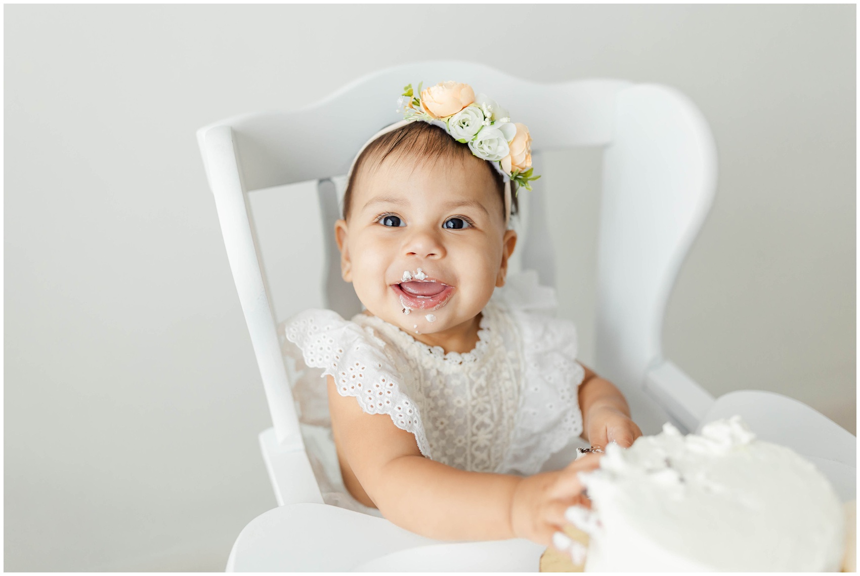 A happy toddler girl in a white dress sits in her high chair covered in cake for her birthday after visiting birthday party venues miami