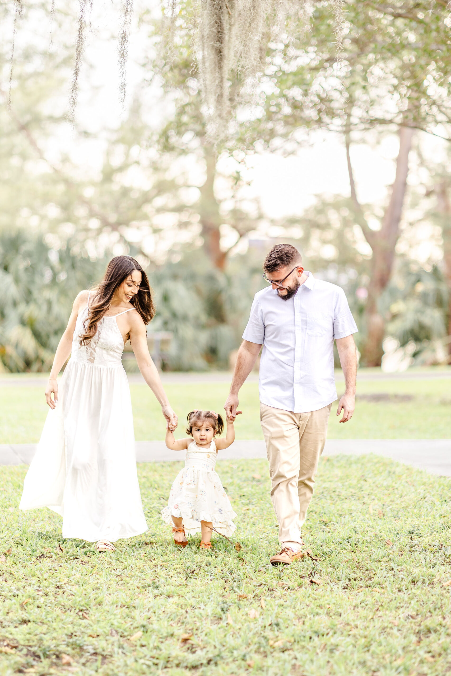 A toddler girl in a yellow dress walks with mom and dad's help through a park