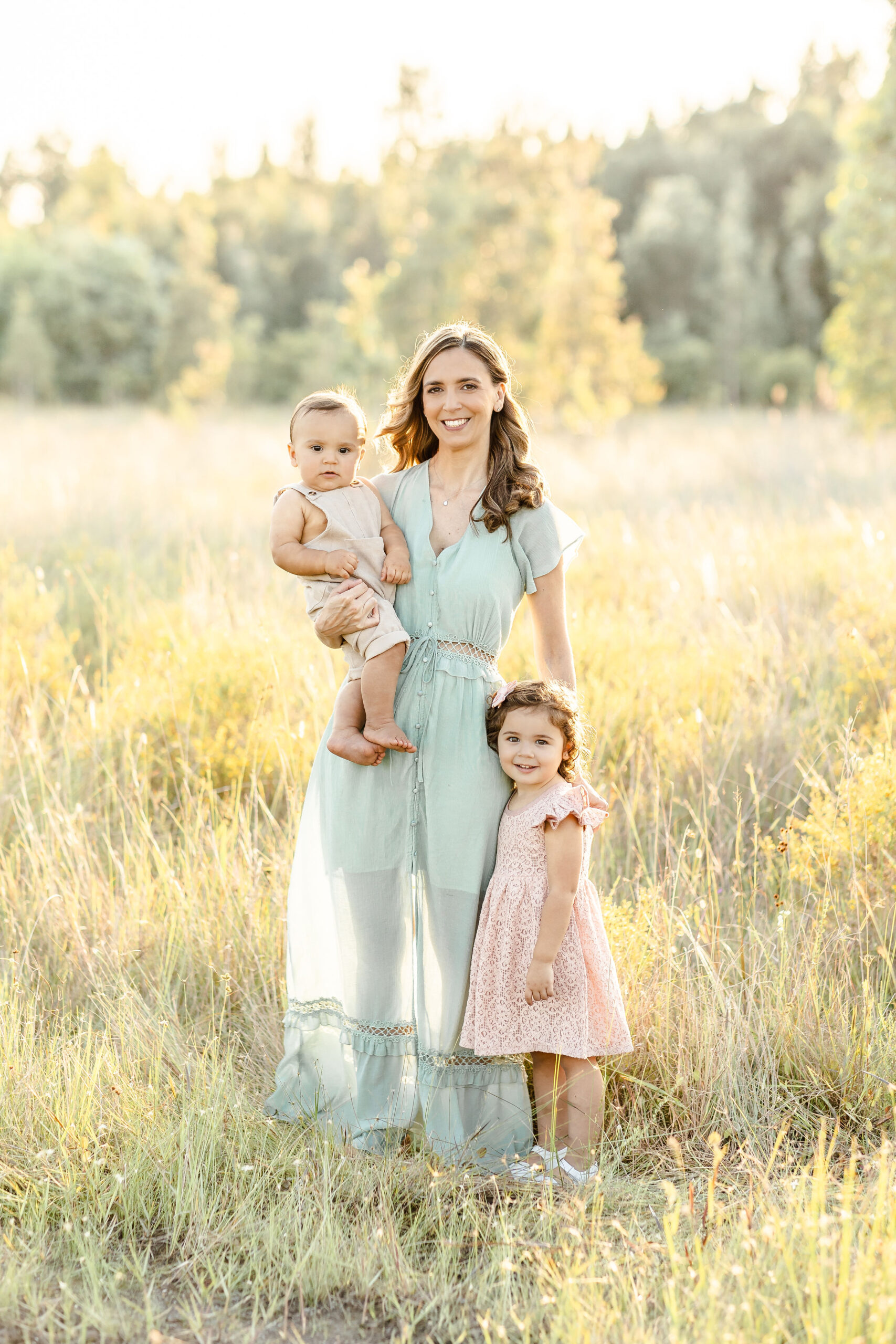 A happy mom stands in a park lawn of tall grass at sunset with her toddler daughter and infant son on her hip before some holiday events miami