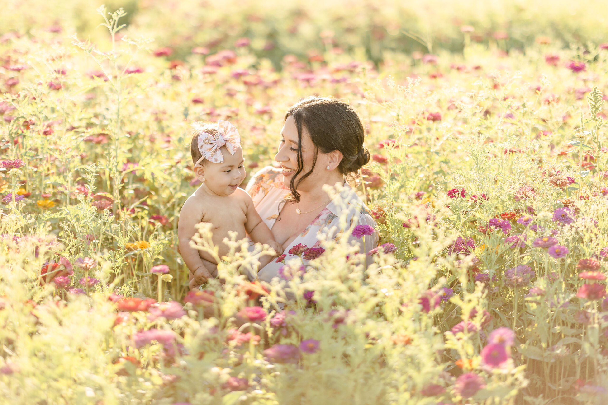 A happy toddler girl in a bow headband sits in mom's lap in a field of colorful wildflowers after visiting miami fall festivals