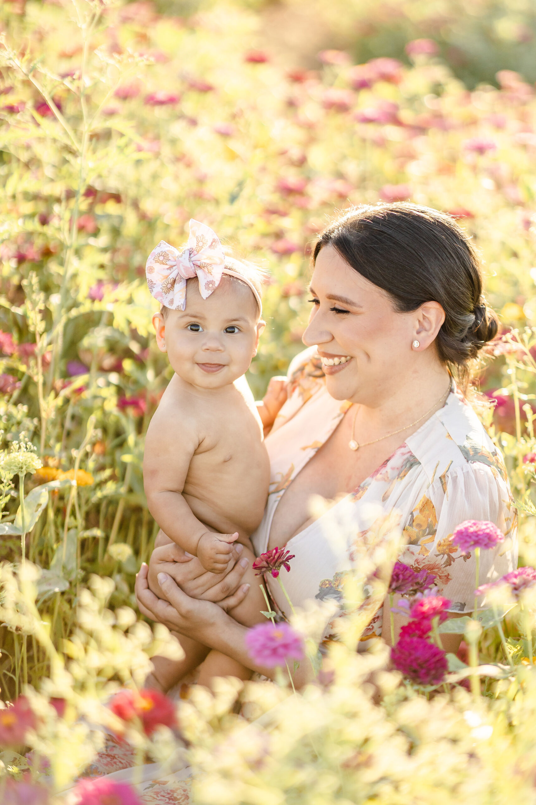 A happy mother smiles with her toddler daughter on her hip while sitting in a field of wildflowers at sunset after visiting miami fall festivals