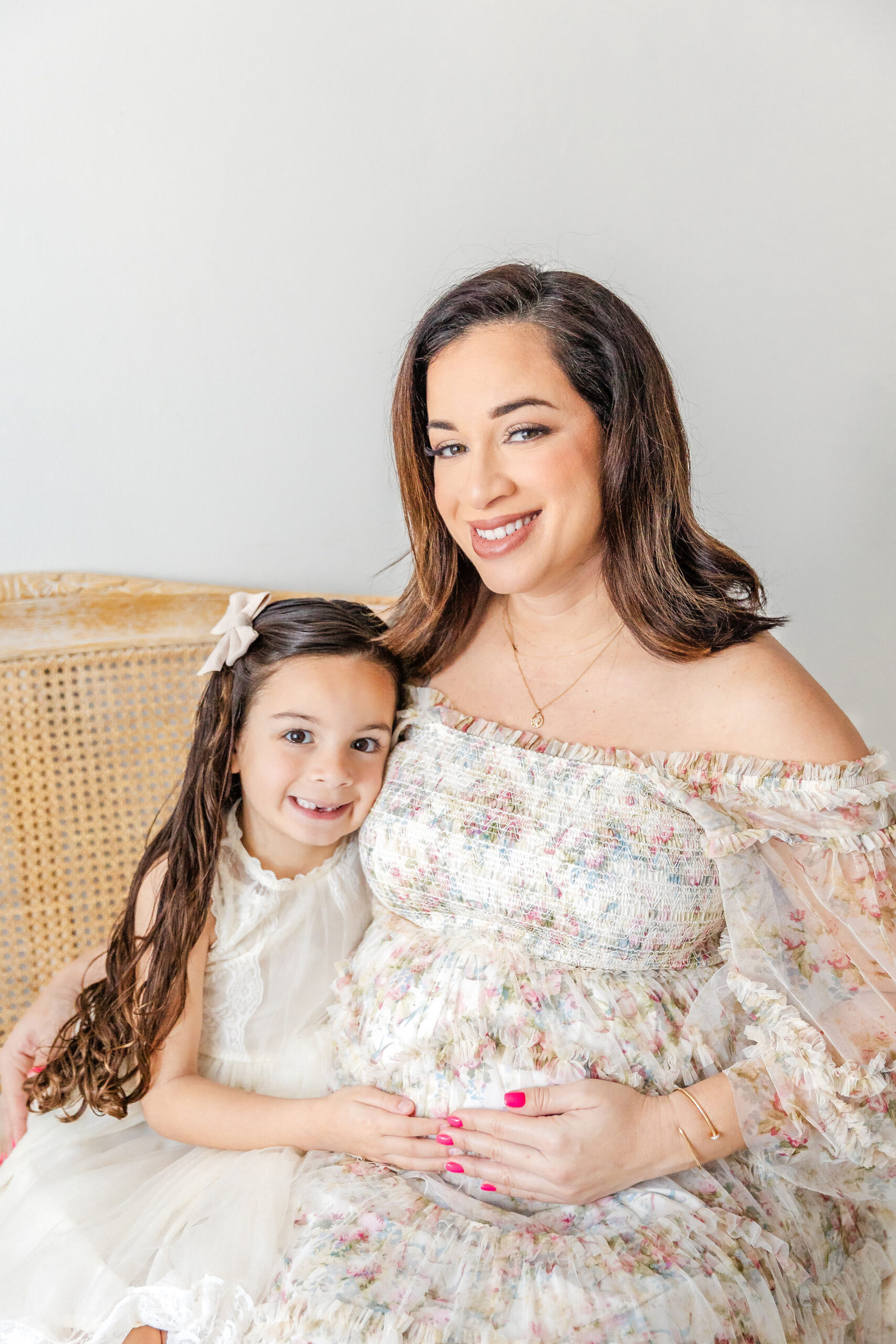 A smiling toddler holds her hand on mom's pregnant bump while sitting with her on a bench in a studio