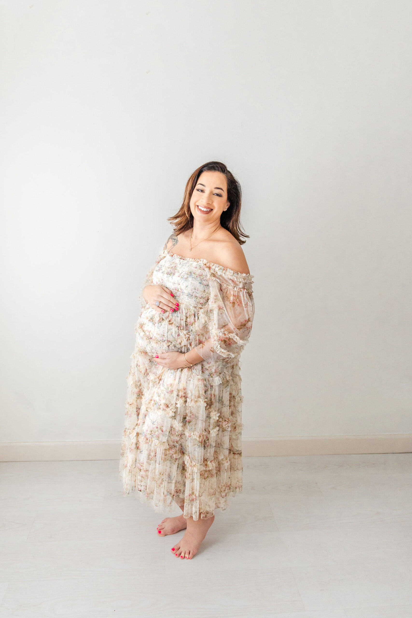 A happy mom to be in a floral print maternity gown stands barefoot in a studio holding her bump after visiting baptist health women's center