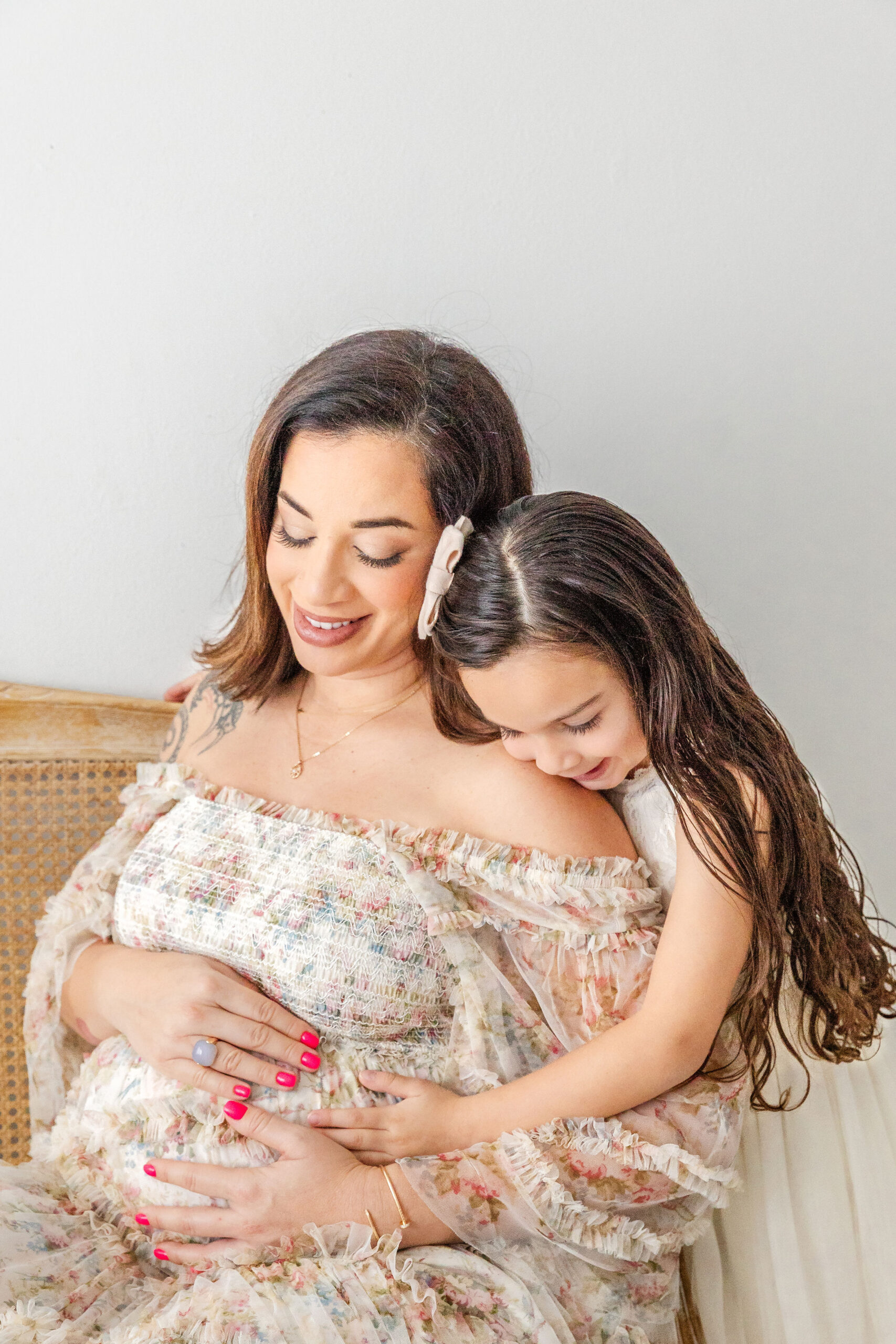 A toddler girl in a white dress hugs onto her pregnant mom while sitting on a bench in a studio holding the bump after visiting baptist health women's center