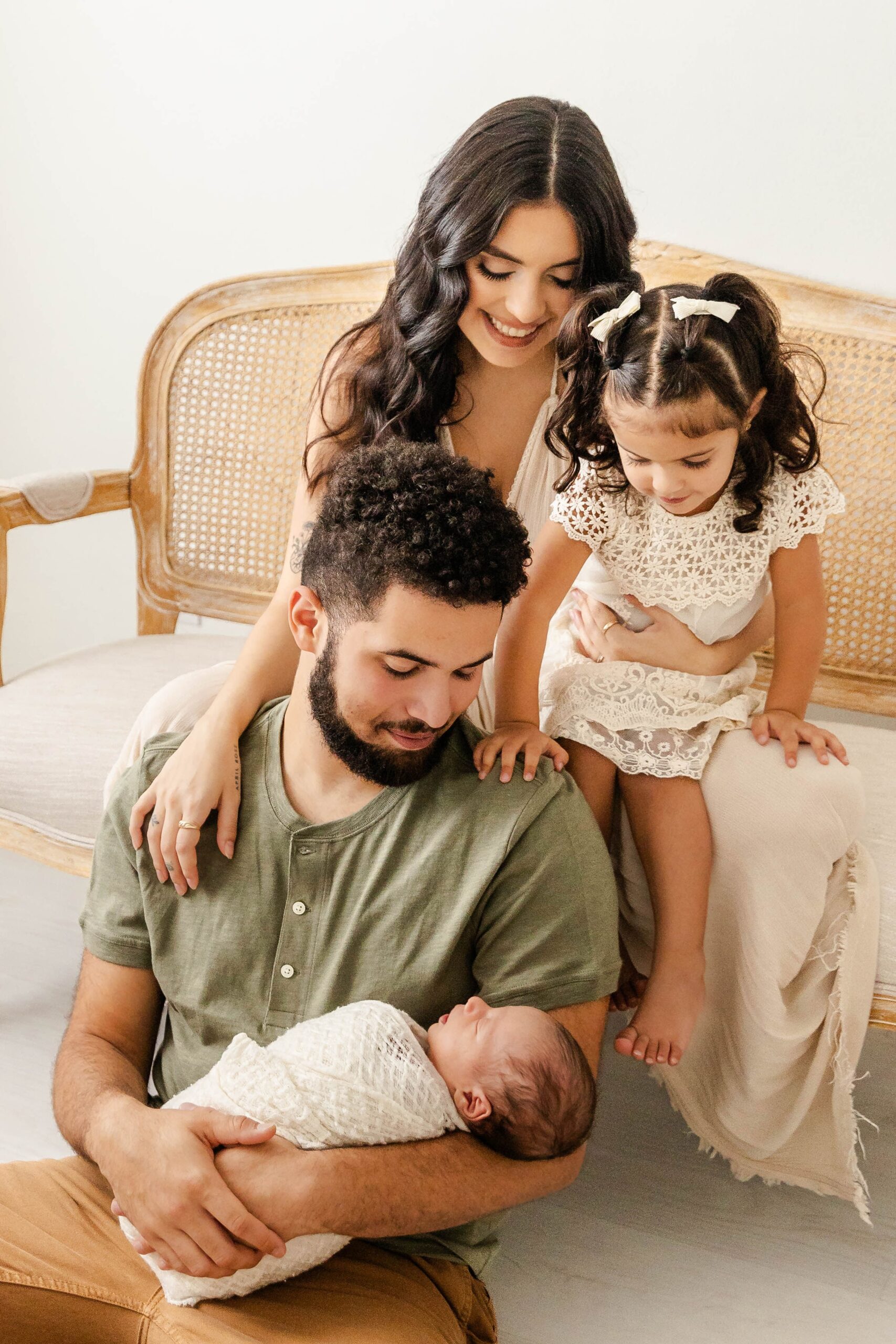 A happy father sits on the floor cradling his newborn baby as mom and toddler daughter sit behind him on a bench