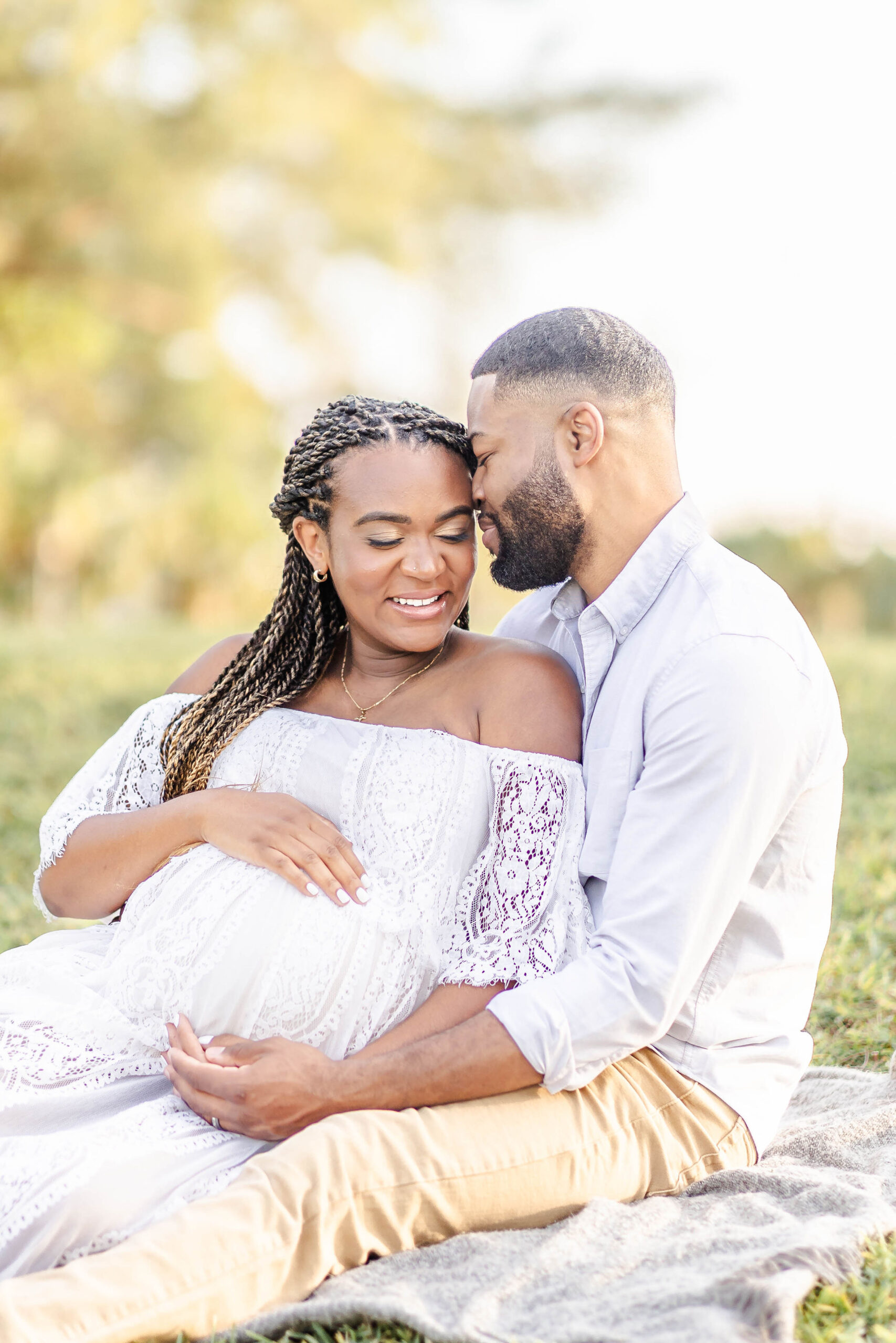 A pregnant woman leans into her husband as they snuggle on a picnic blanket in a park at sunset