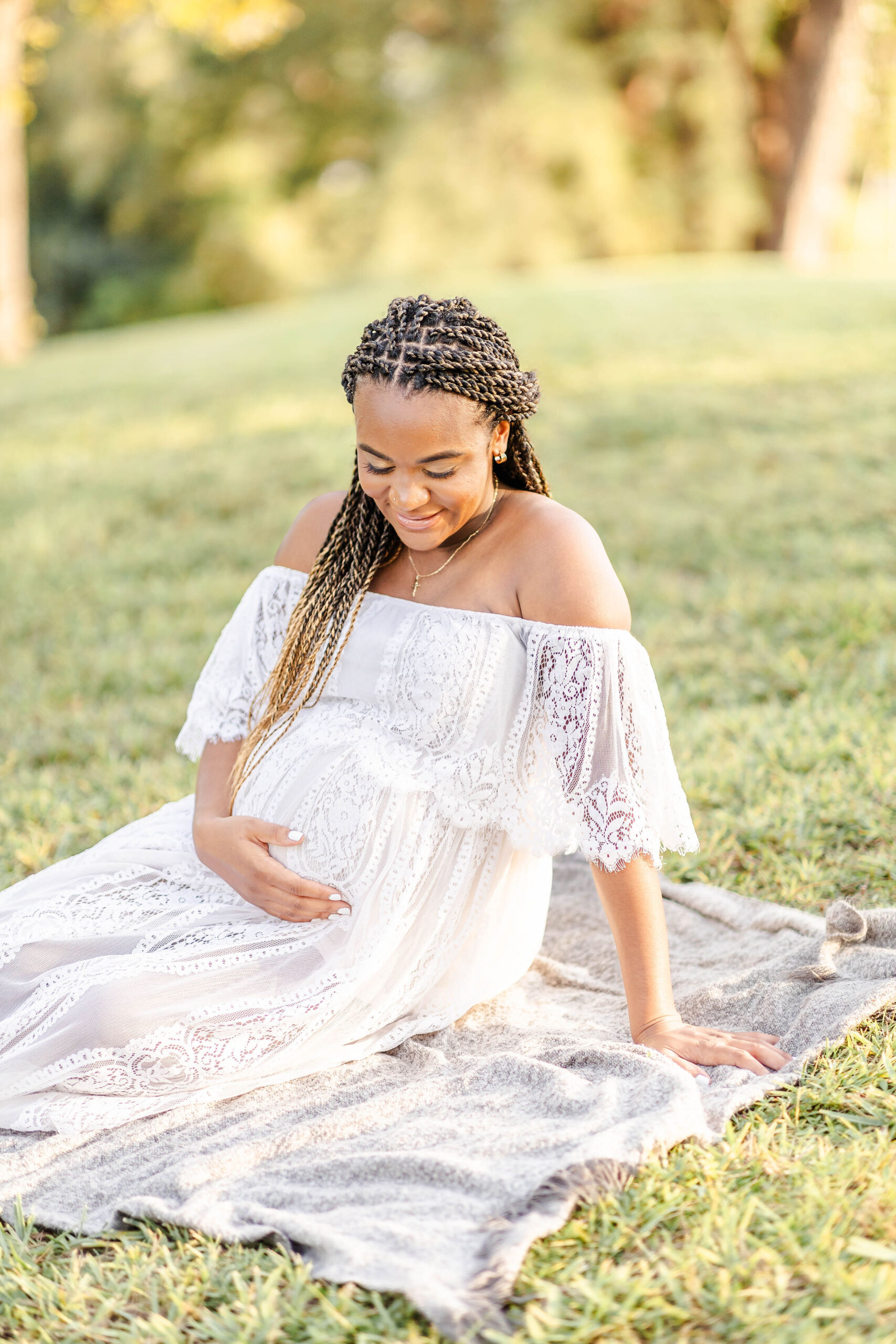 A smiling mom to be sits on a picnic blanket in a park lawn smiling down at her bump in a white lace maternity gown after visiting mercy hospital obgyn