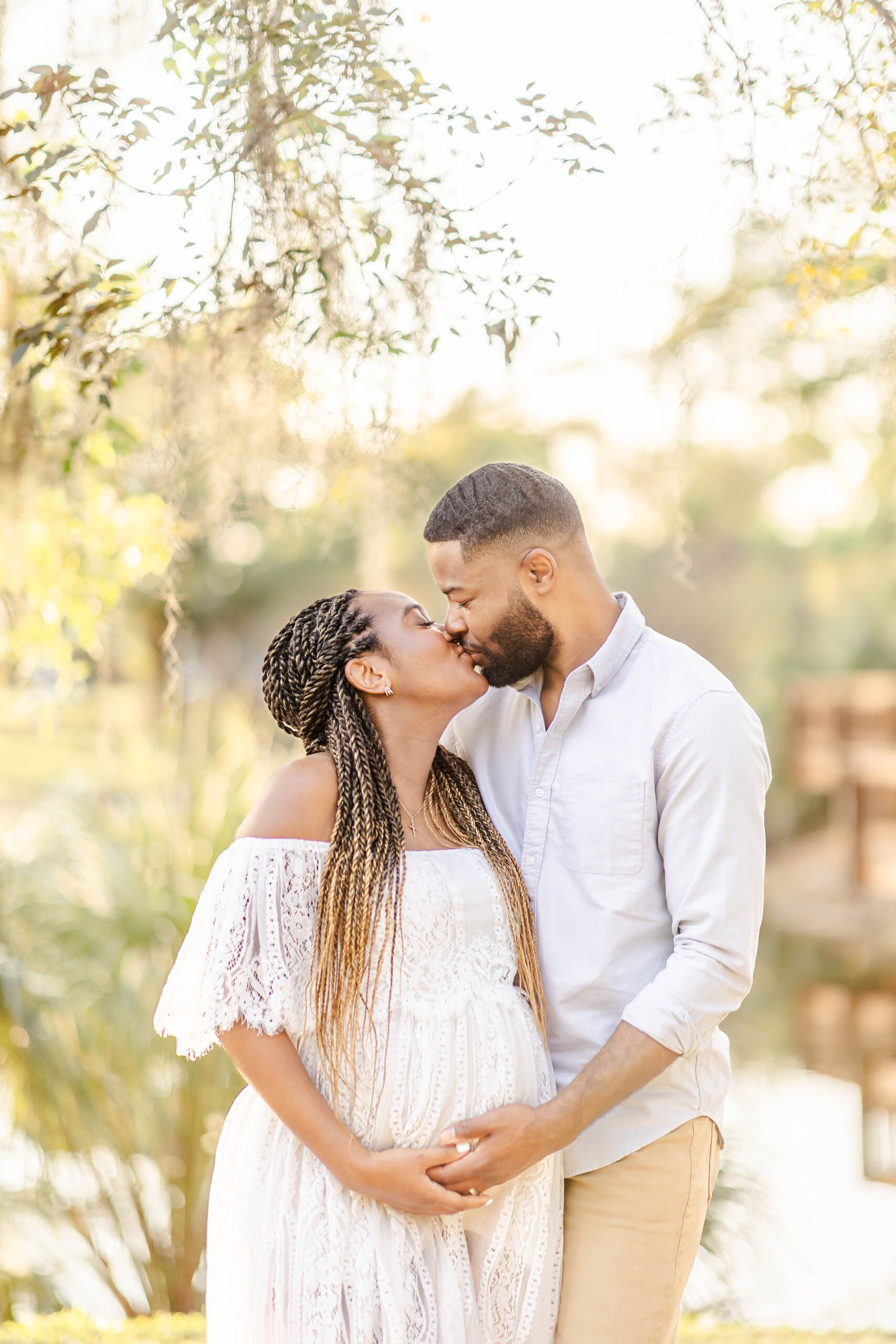 Expecting parents stand by a pond under some trees kissing and holding the bump after visiting mercy hospital obgyn