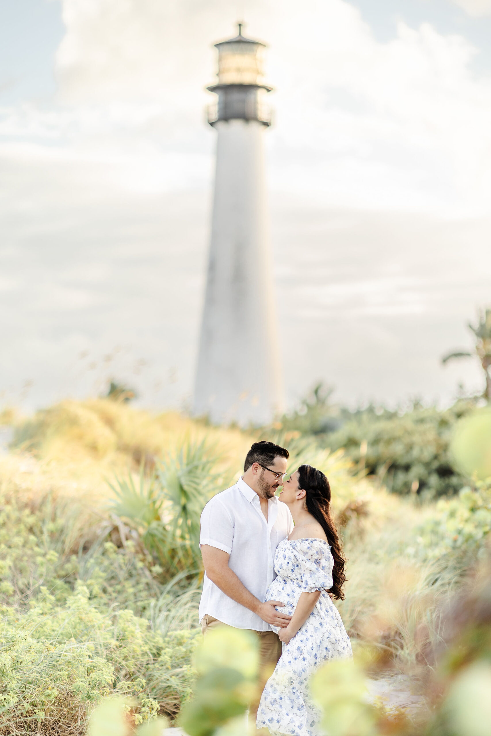 Mom and dad to be stand in a beach dune kisses beneath the lighthouse at sunset