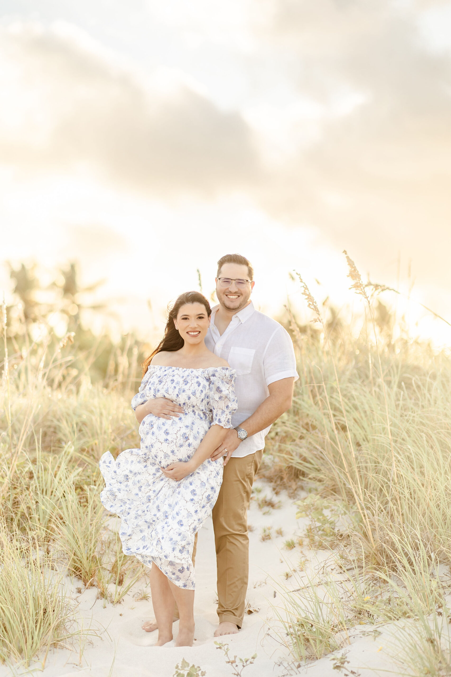 Happy expecting parents stand in a beach dune while holding the bump at sunset after visiting obgyn university of miami