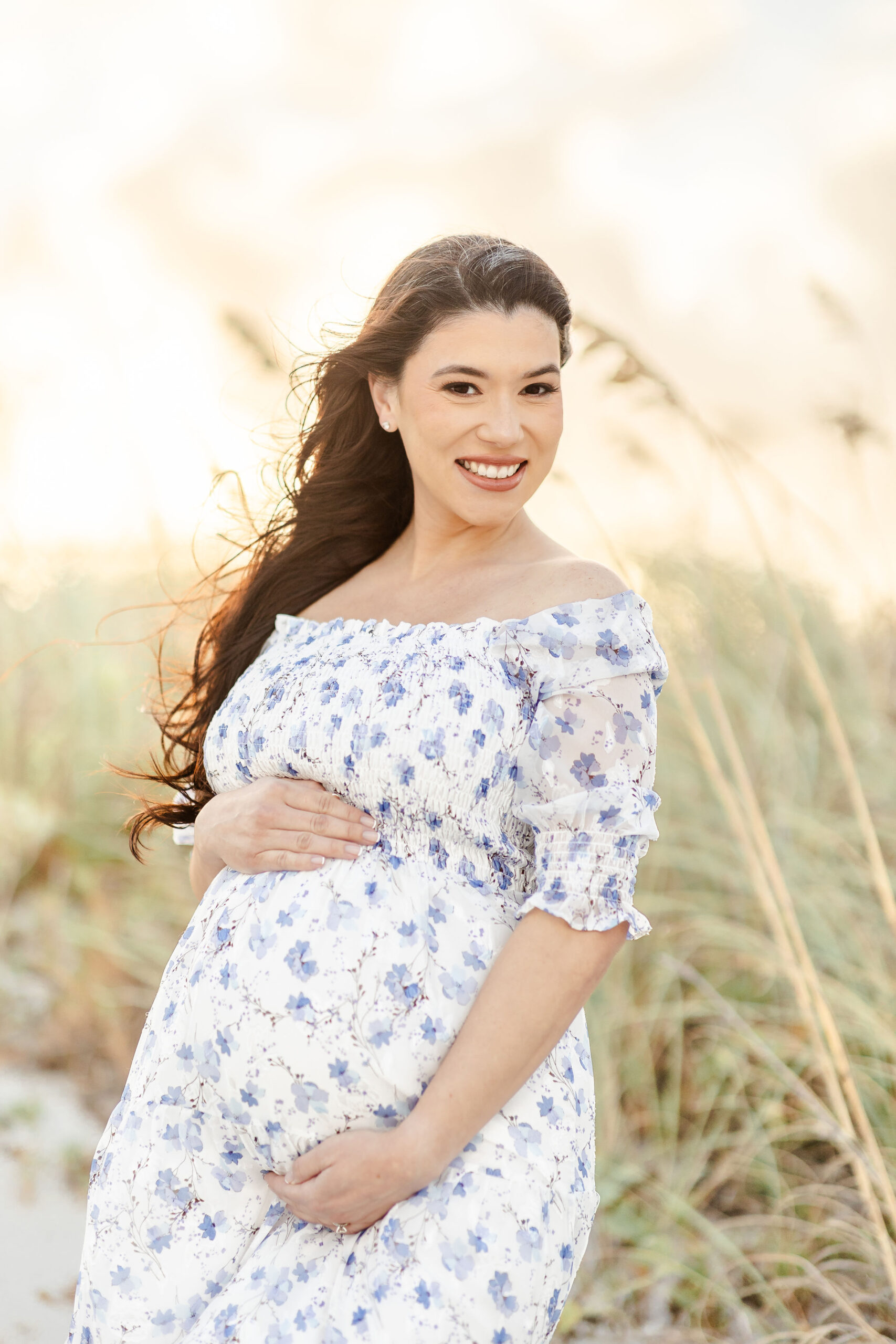 A smiling mom to be in a blue floral print maternity dress stands on a windy beach at sunset after visiting obgyn university of miami