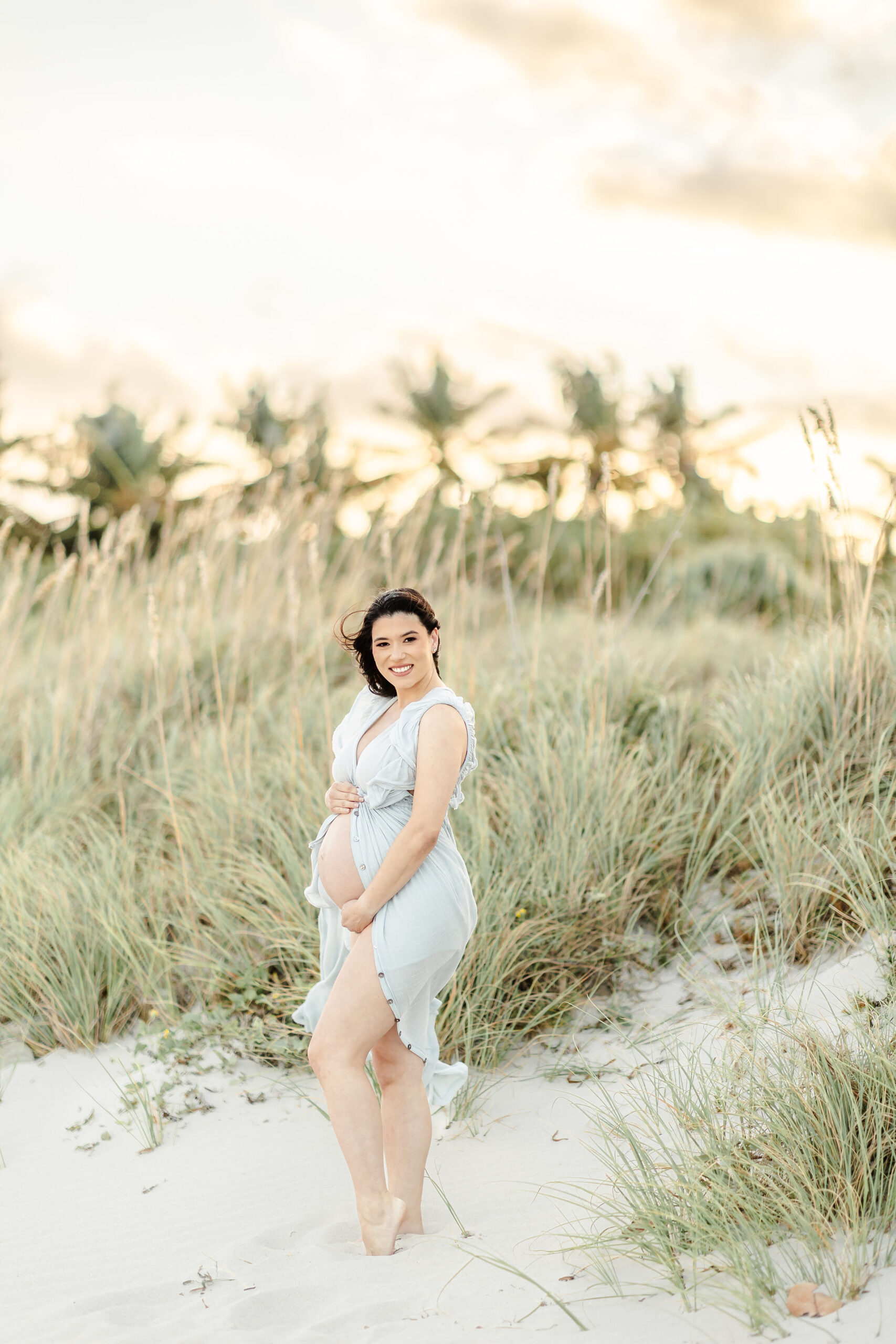 A happy pregnant woman with her dress open to show her bump stands in a beach dune at sunset