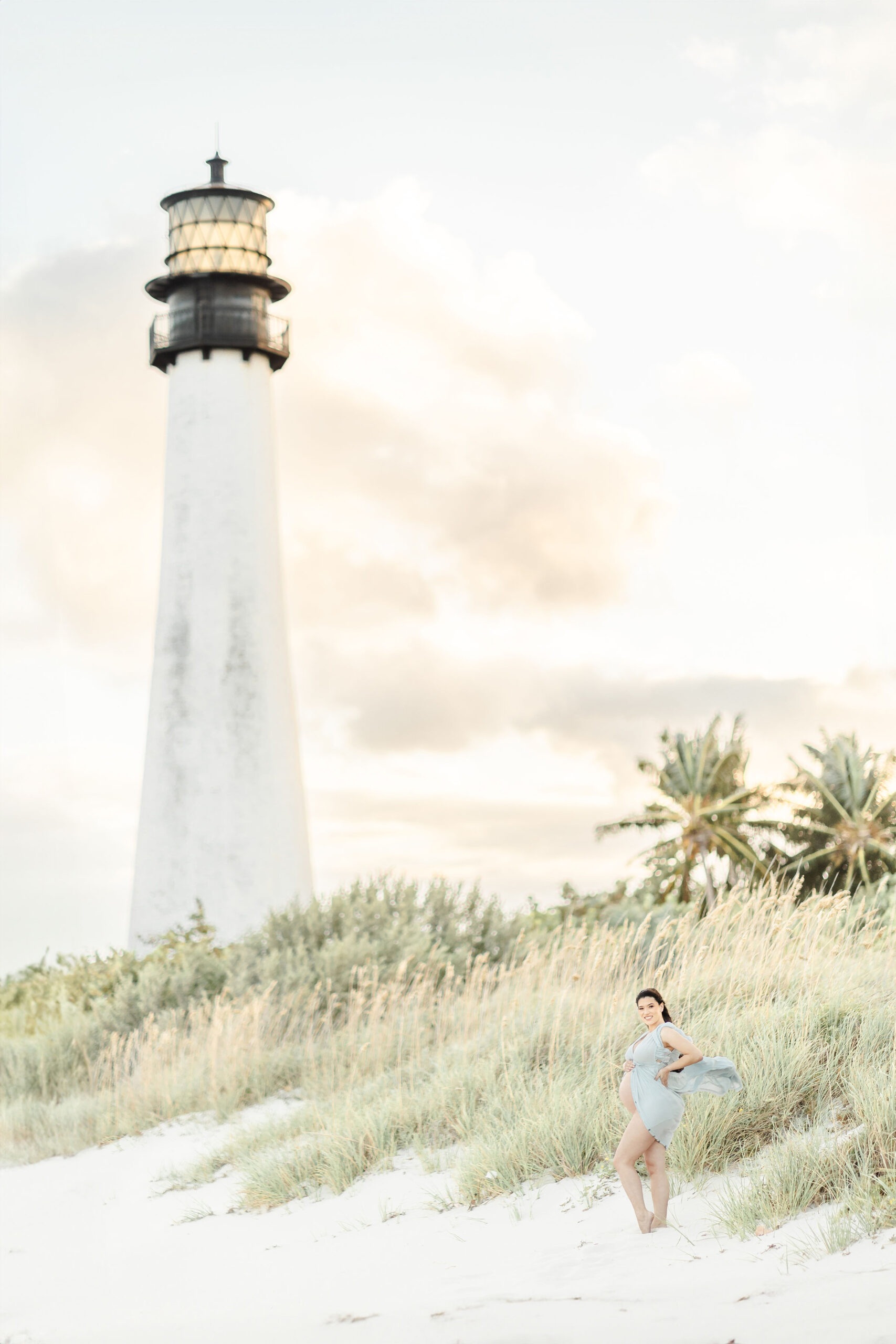 A mom to be stands with her bump out on the beach dune under a lighthouse after visiting Macanoco & Co.