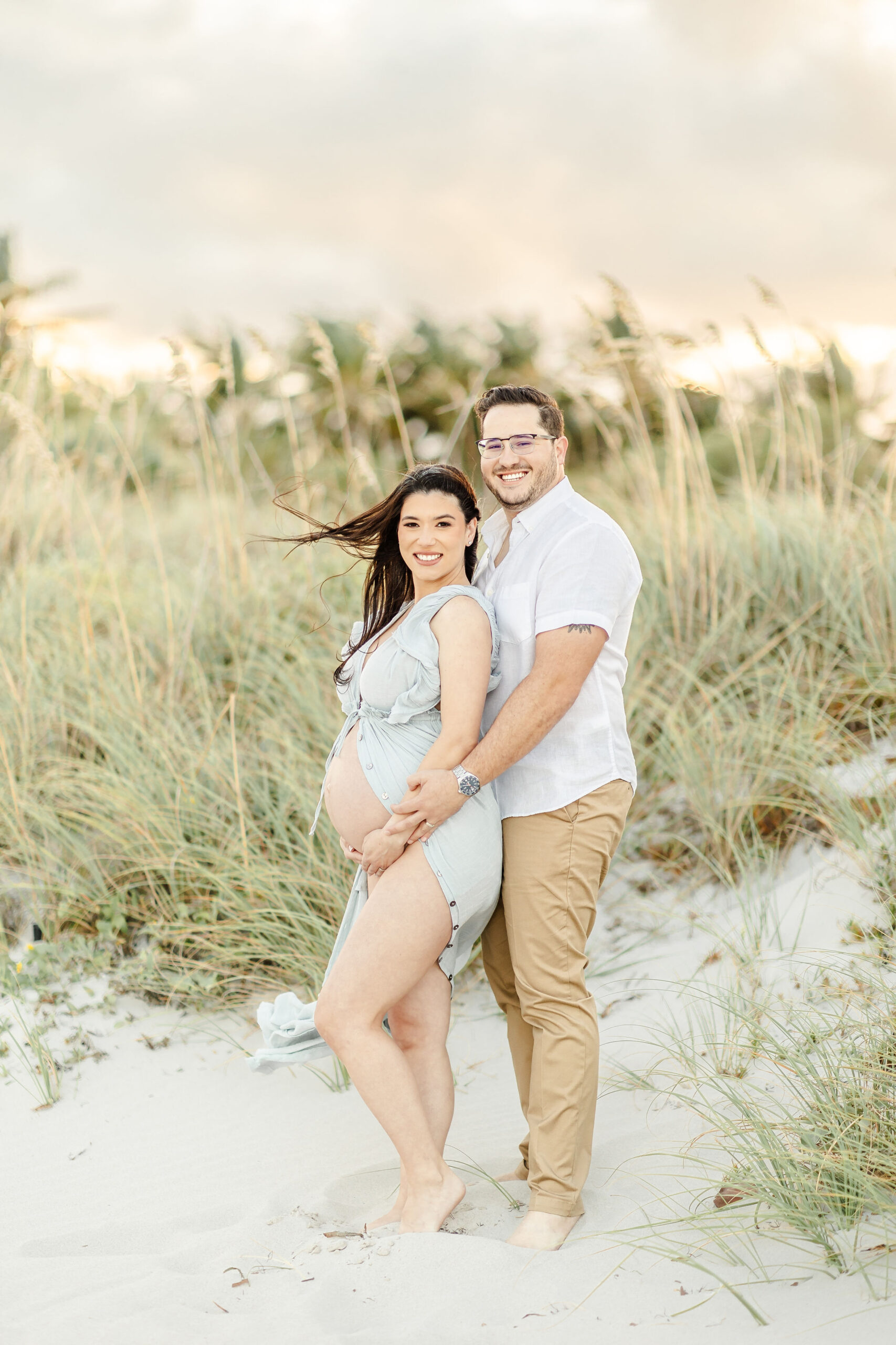 Smiling expecting parents stand in the beach dune with hands on the exposed bump on a windy day before visiting Macanoco & Co.