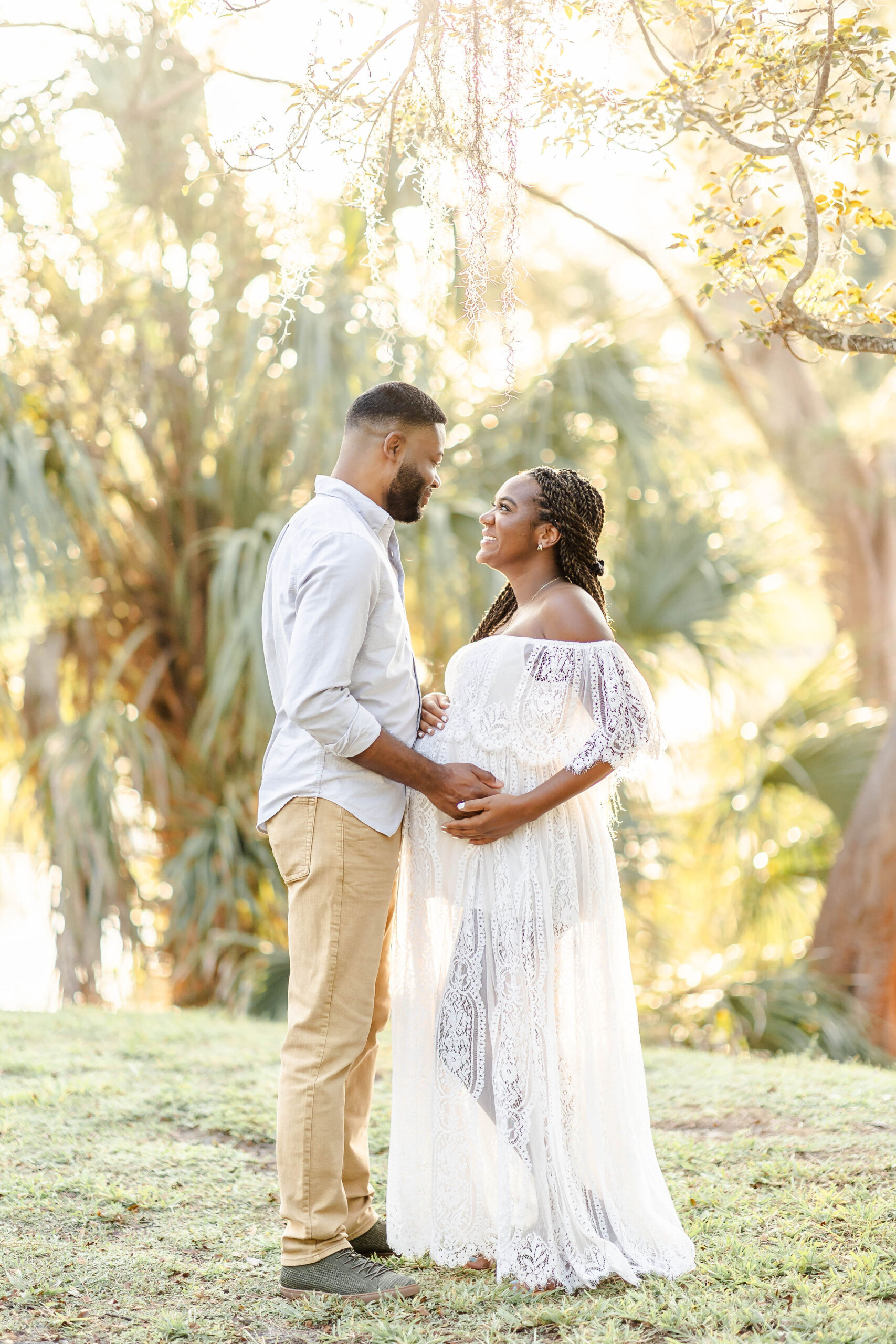 Happy expecting couple smiling at each other while standing in a lawn with hands on bump