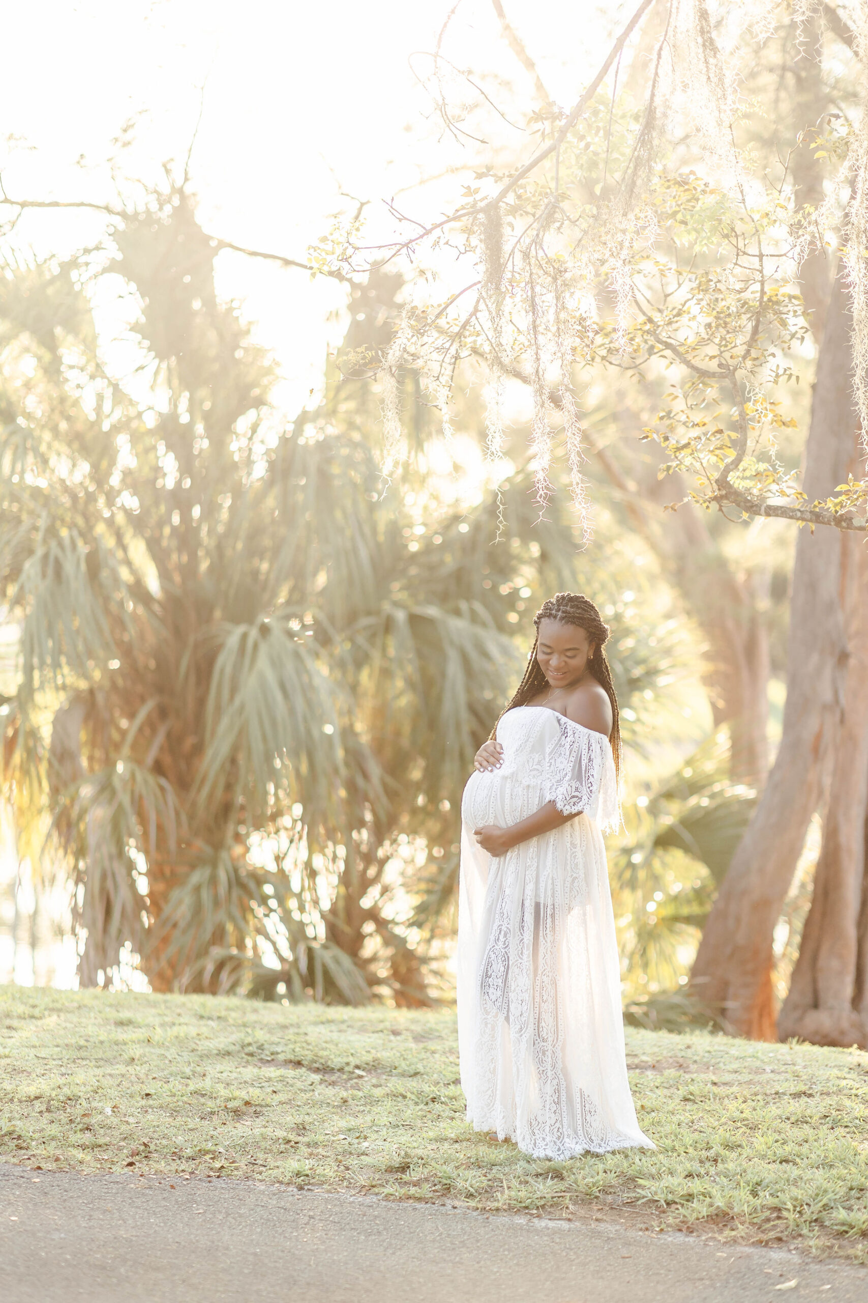 A mom to be in a lace white maternity gown smiles down to her bump in her hands while standing in the sunshine in a park after visiting Midwife and Co Miami