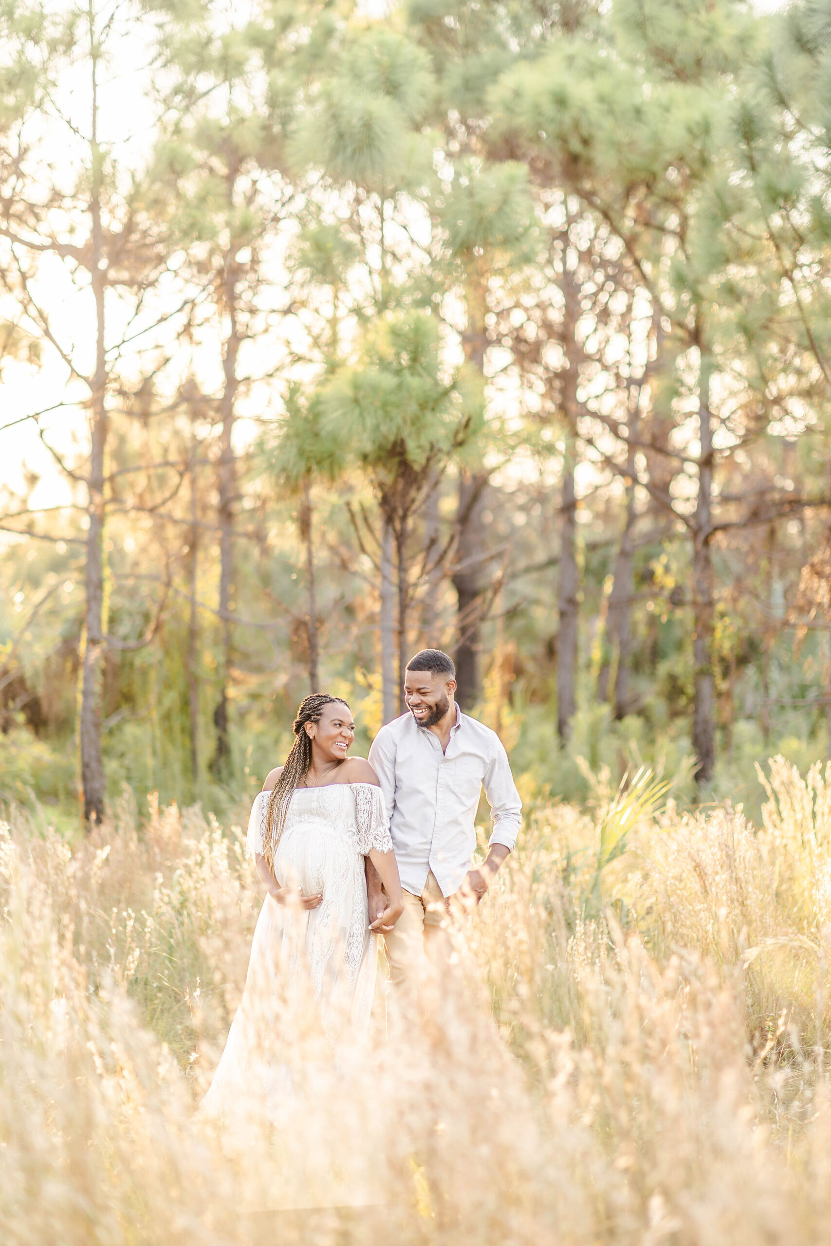 Expecting parents laugh while exploring tall grasses in white at sunset after meeting Midwife and Co Miami