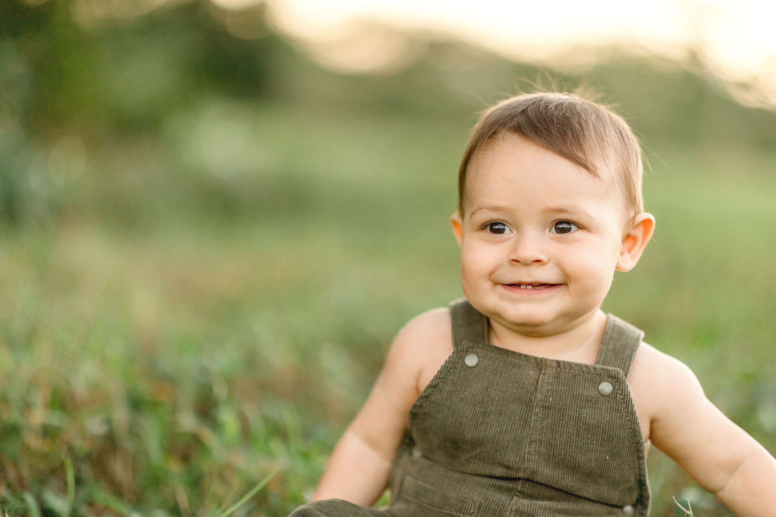 A smiling baby with a couple teeth sits in a lawn at sunset in green overalls