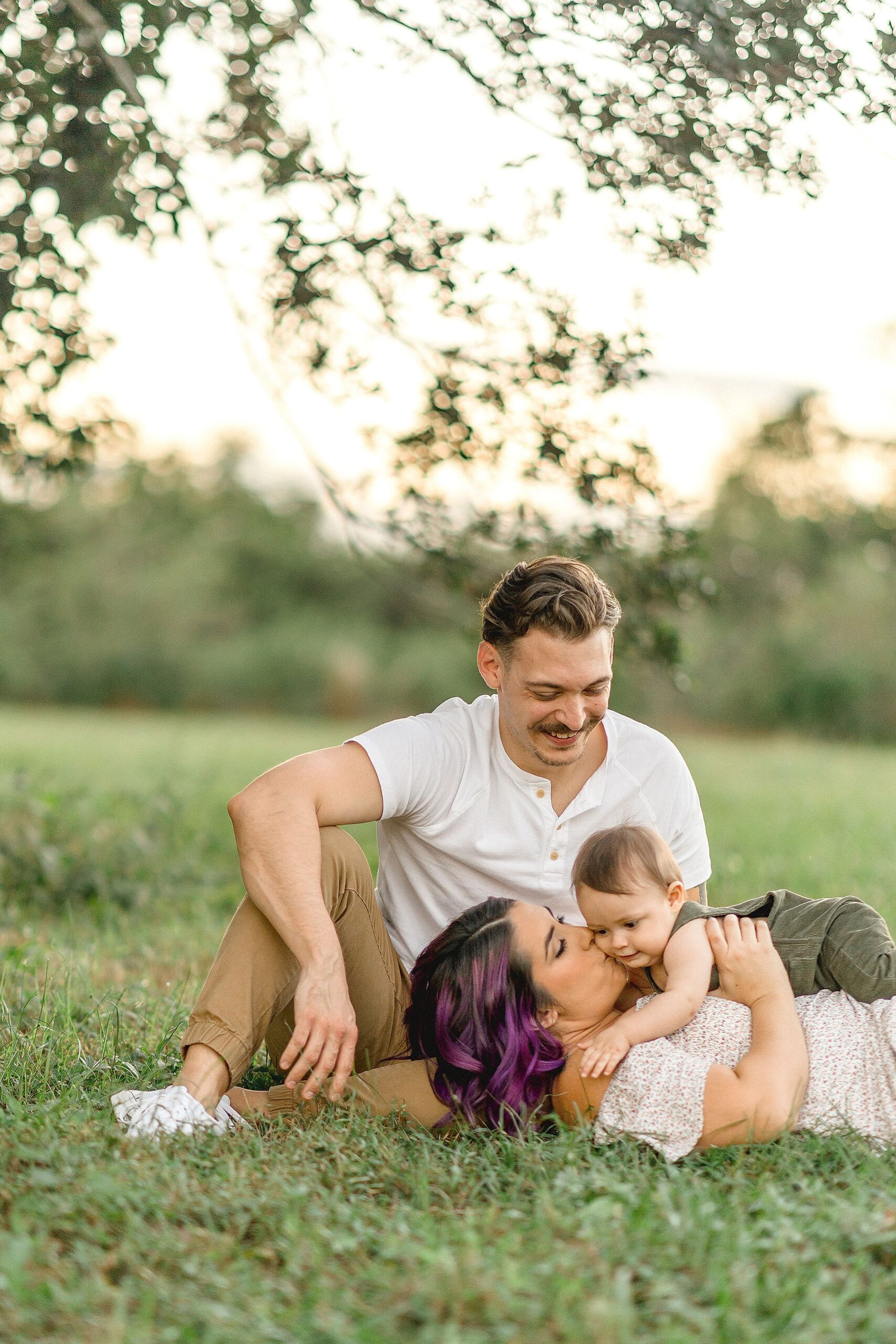 A happy dad looks on as his wife kisses their baby while laying in the grass at sunset after visiting coral gables family chiropractic