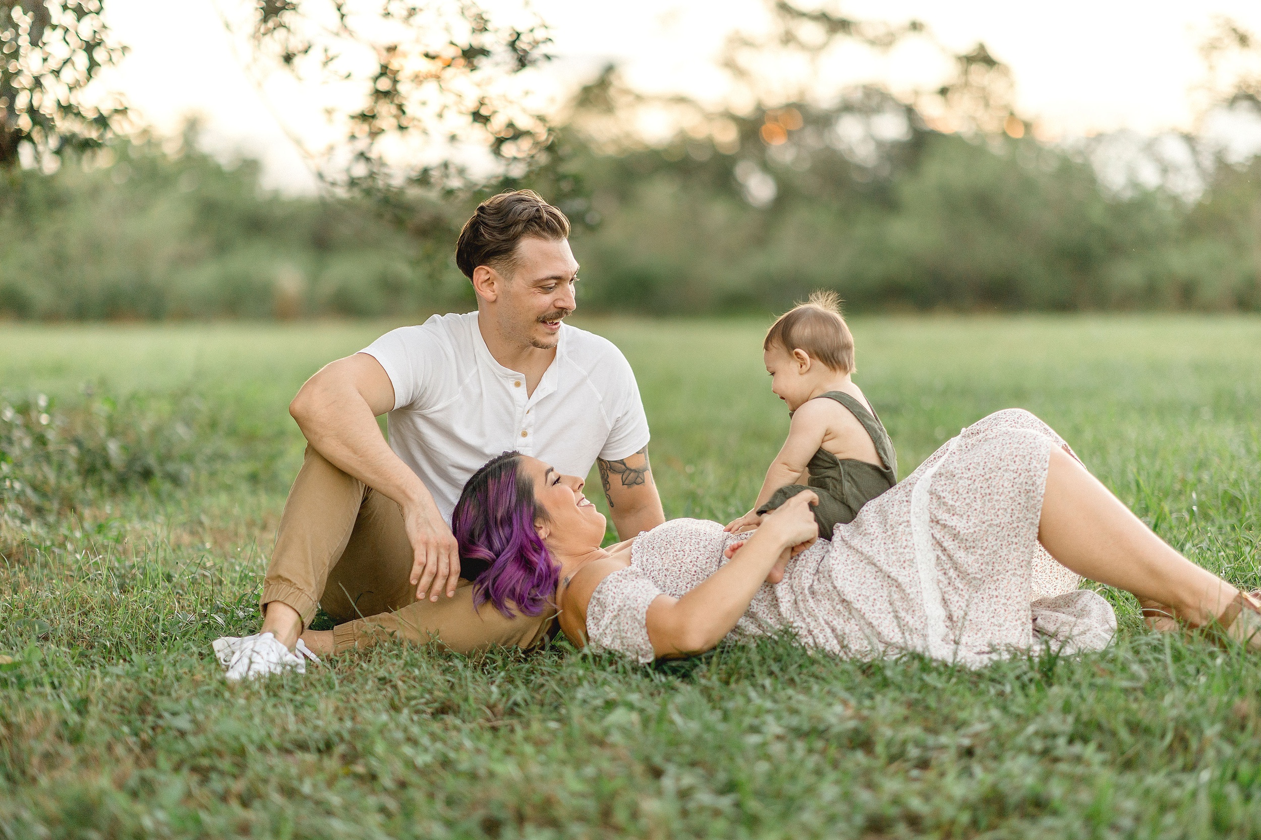 Happy mom and dad play with their baby on mom's stomach while laying in a park lawn at sunset after visiting coral gables family chiropractic