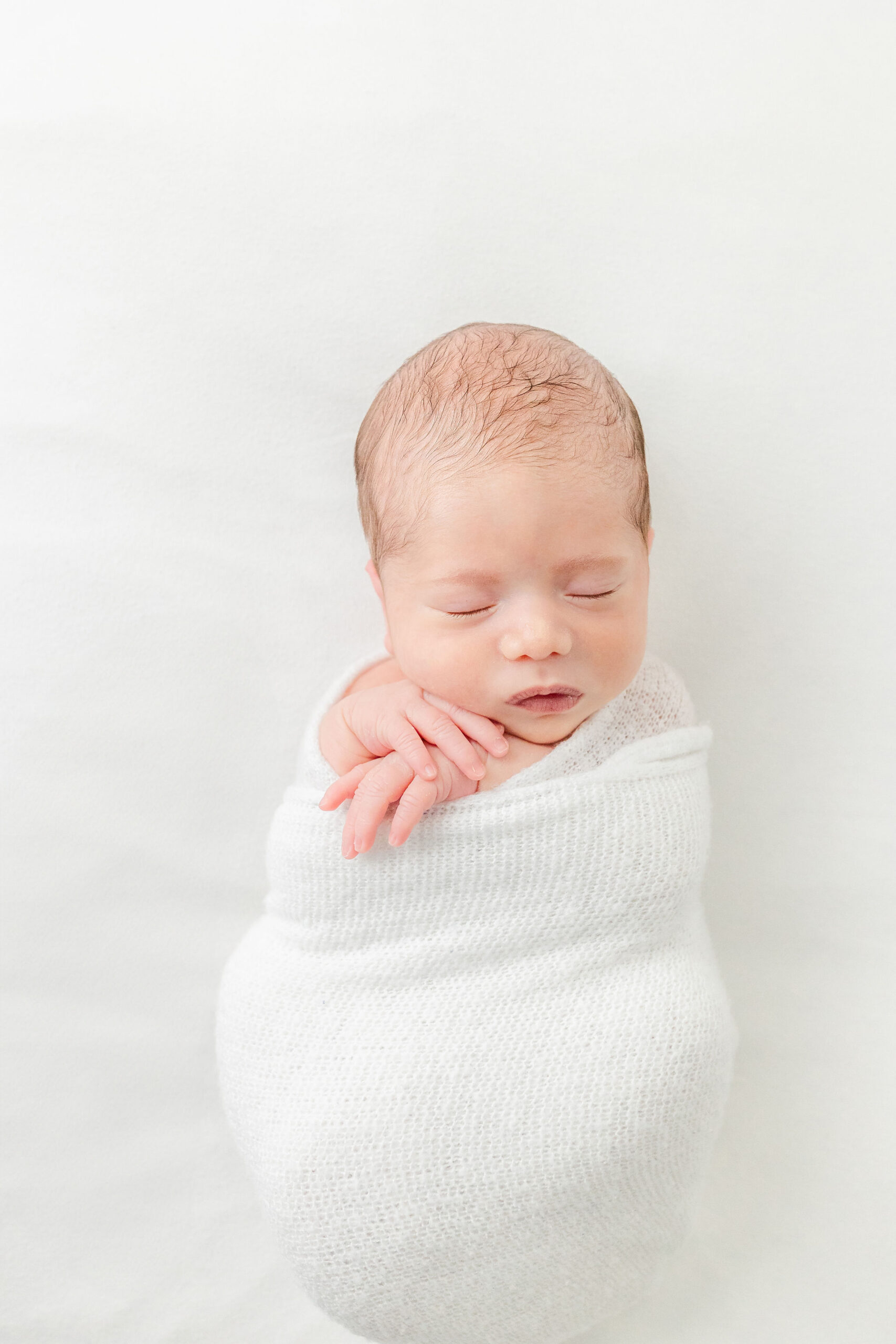 A newborn baby sleeps in a tight white swaddle in a studio