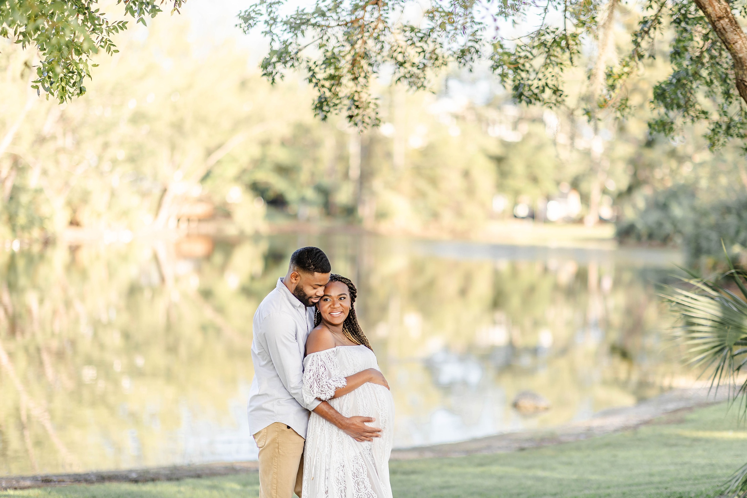 A man hugs his pregnant wife as she stands by a pond in a white lace maternity gown