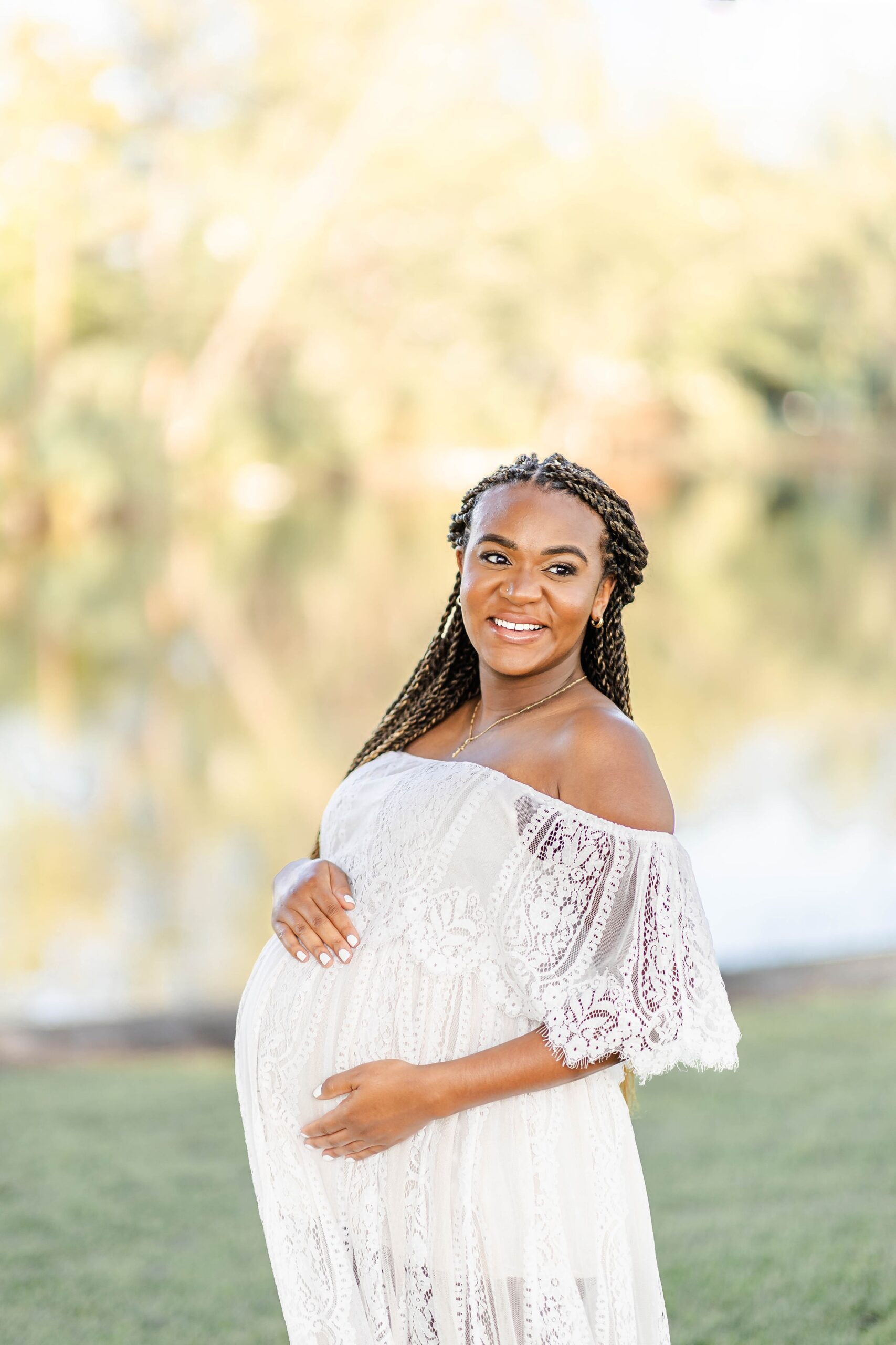 A smiling mom to be in a white lace maternity gown stands in a field at sunset after meeting blooming bellies