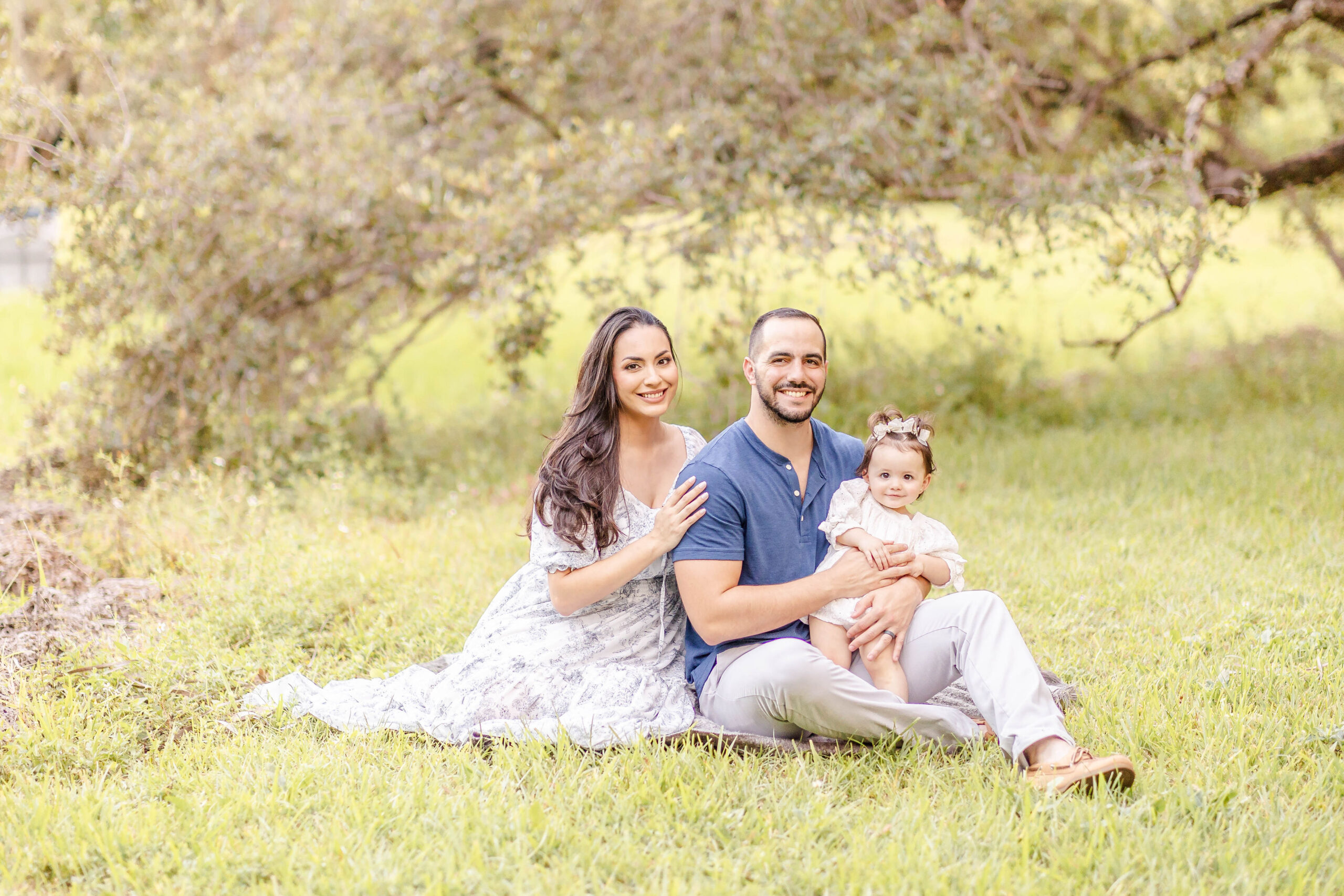 Happy parents smile while sitting in a park lawn with their baby daughter in dad's lap