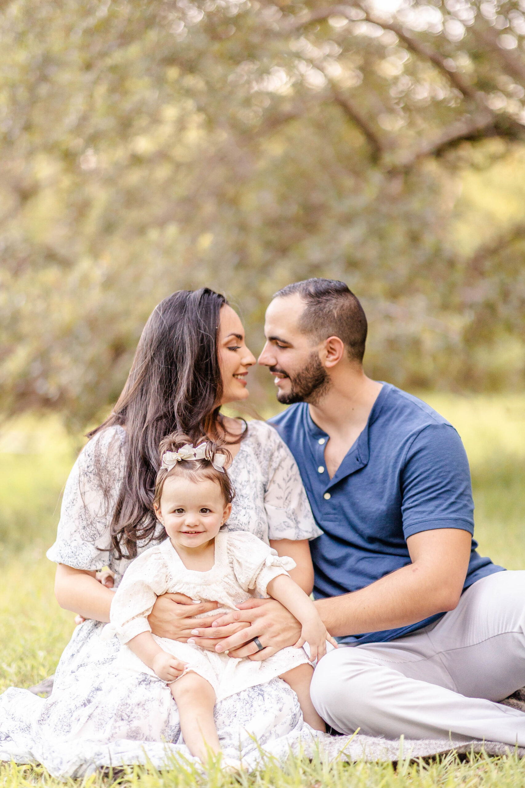 Happy mom and dad sit on a blanket in a park touching noses with their baby daughter in a dress in mom's lap after meeting coral gables pediatricians