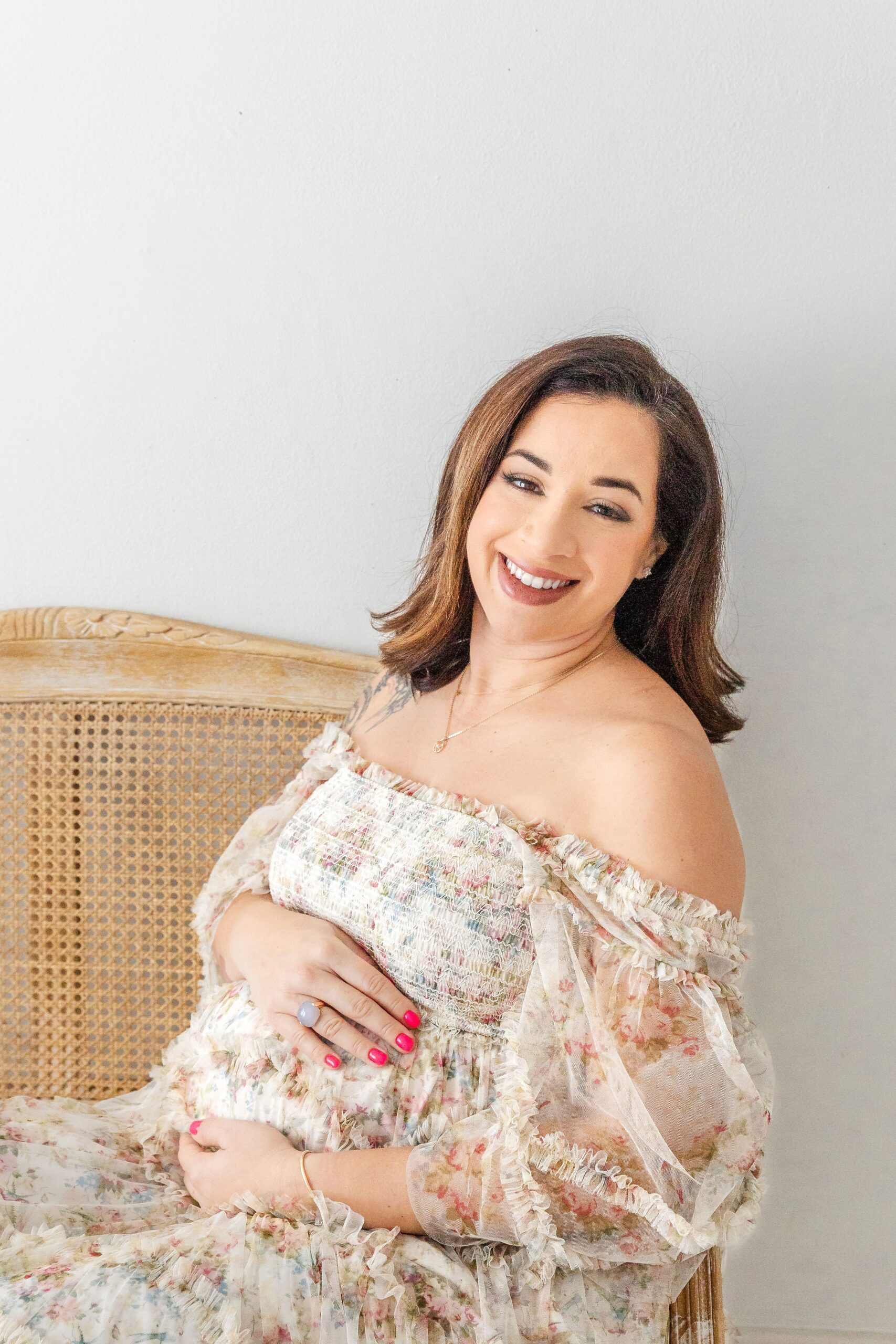 A smiling mother to be in a floral print maternity gown sitting on a bench in a studio