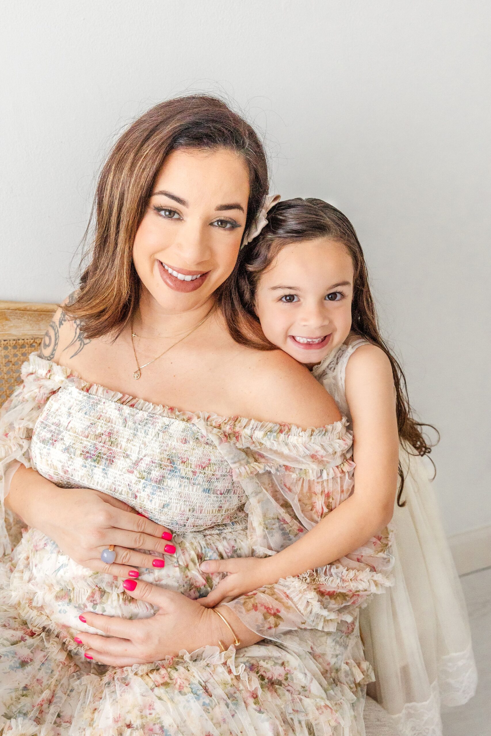 A toddler girl hugs her pregnant mom while sitting on a bench in a studio after visiting hello baby ultrasound miami