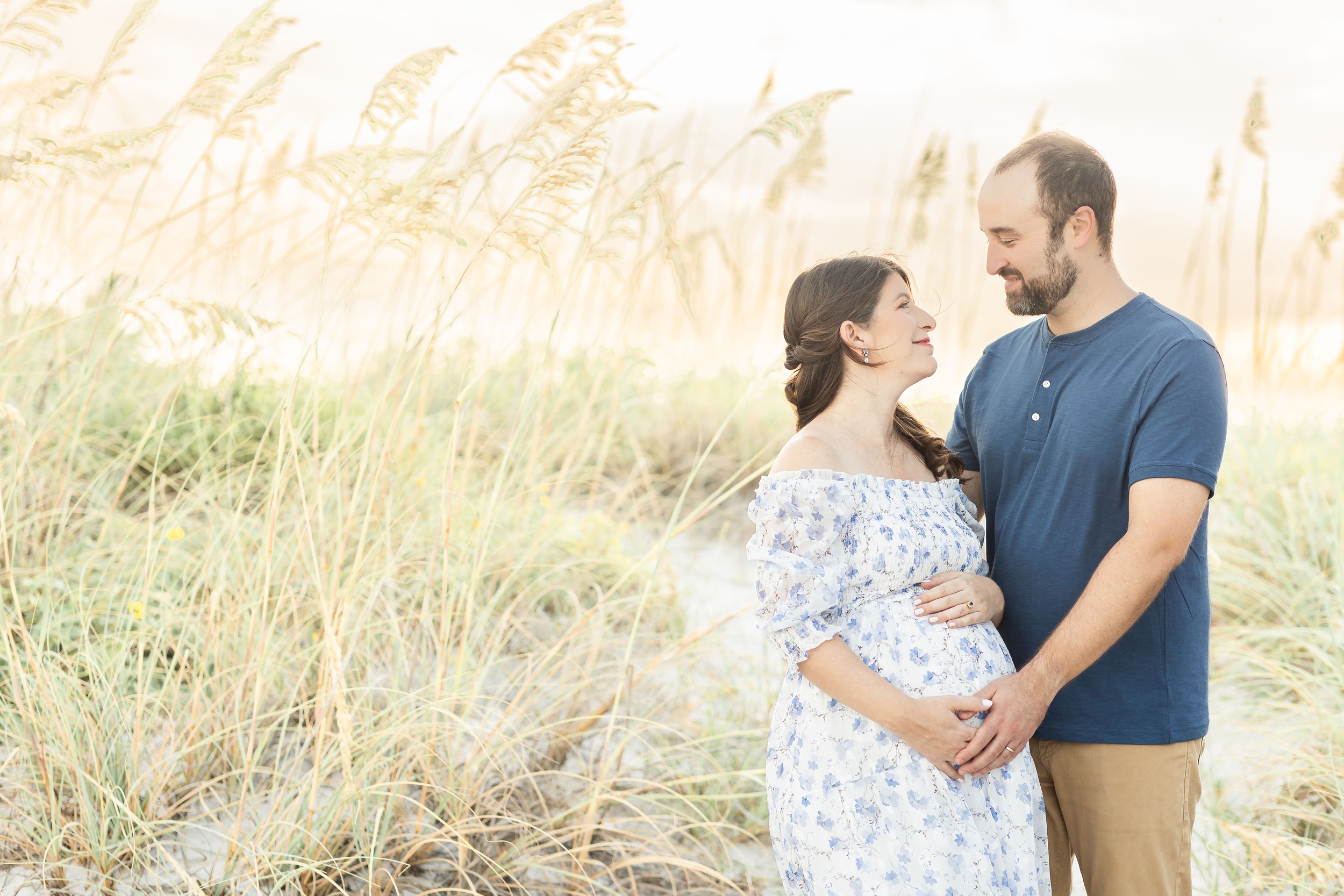 A happy pregnant couple smiles at each other while holding the bump in a golden beach dune at sunset