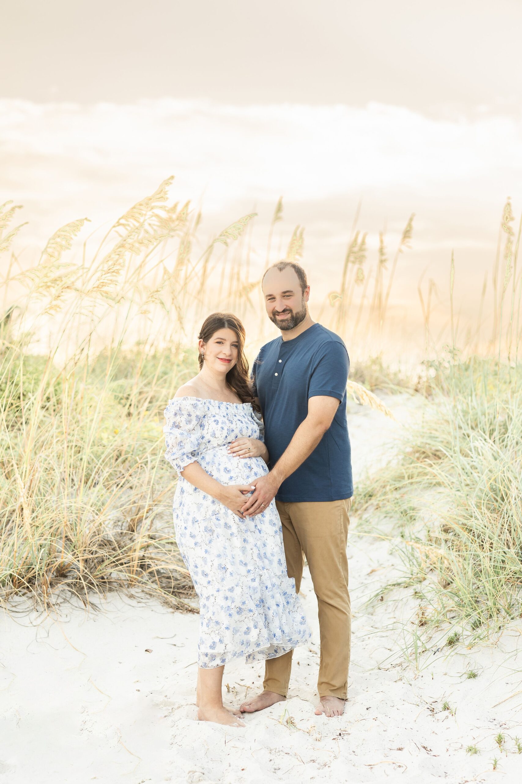 A smiling pregnant couple touches the bump while standing on a beach dune at sunset after visiting painless pregnancy