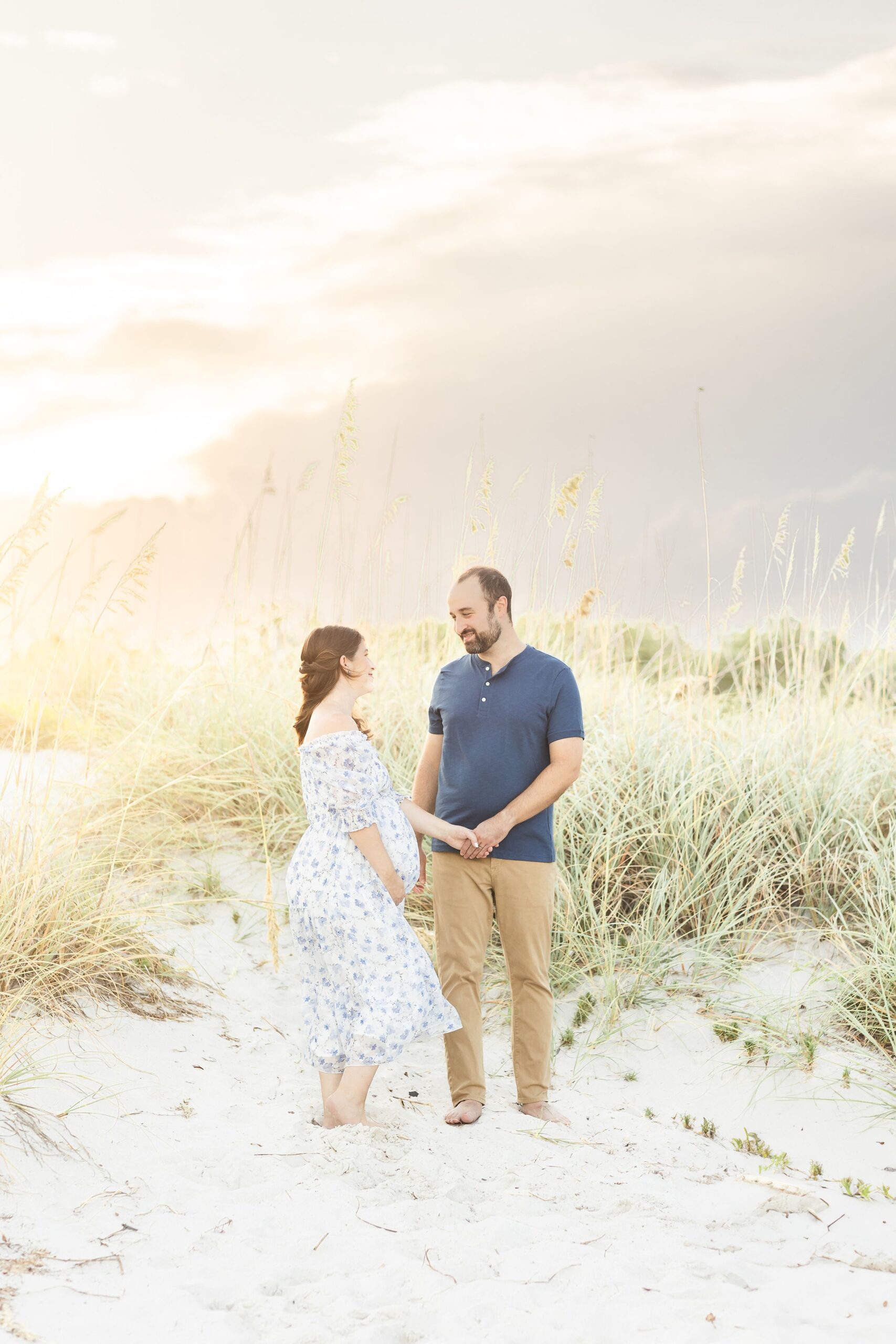 A happy expecting couple shares a smile while holding hands on a beach dune at sunset after meeting painless pregnancy