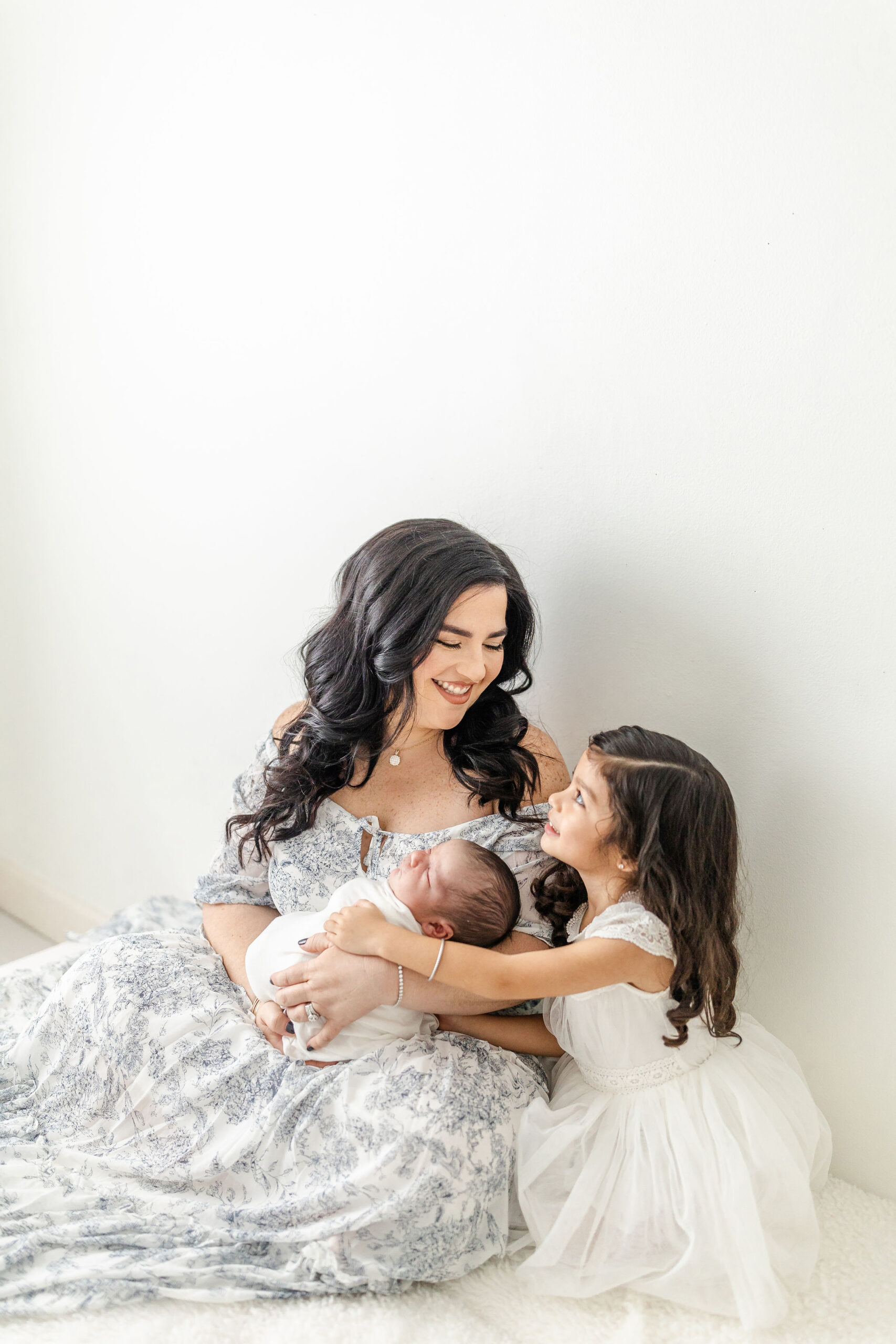 A toddler girl in a white dress hugs her sleeping newborn sibling while sitting with mom against a wall after meeting a postpartum doula in miami